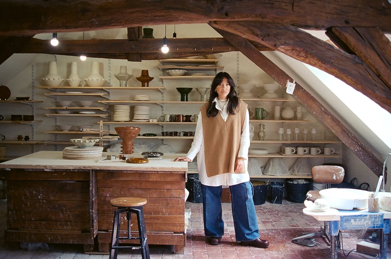 Femme dans un atelier de poterie, entourée de vases, bols, et autres objets en céramique, sous une charpente en bois.
