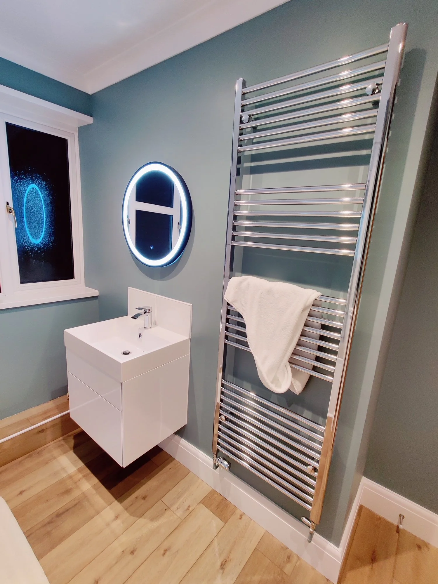 Modern bathroom with a white wall-mounted sink, an illuminated circular mirror, a chrome heated towel rail with a white towel, a window with a frosted glass reflection, and wooden flooring.