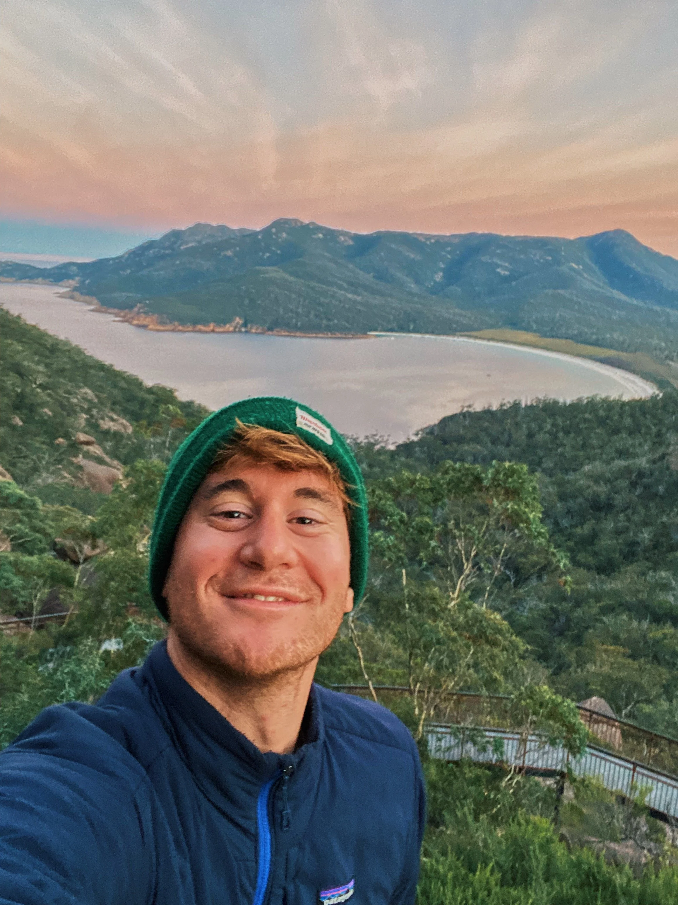 een jonge man neemt een selfie op een uitkijkpunt met een groot meer en bergen op de achtergrond tijdens zonsopgang, hij draagt een groene muts en een blauwe jas.