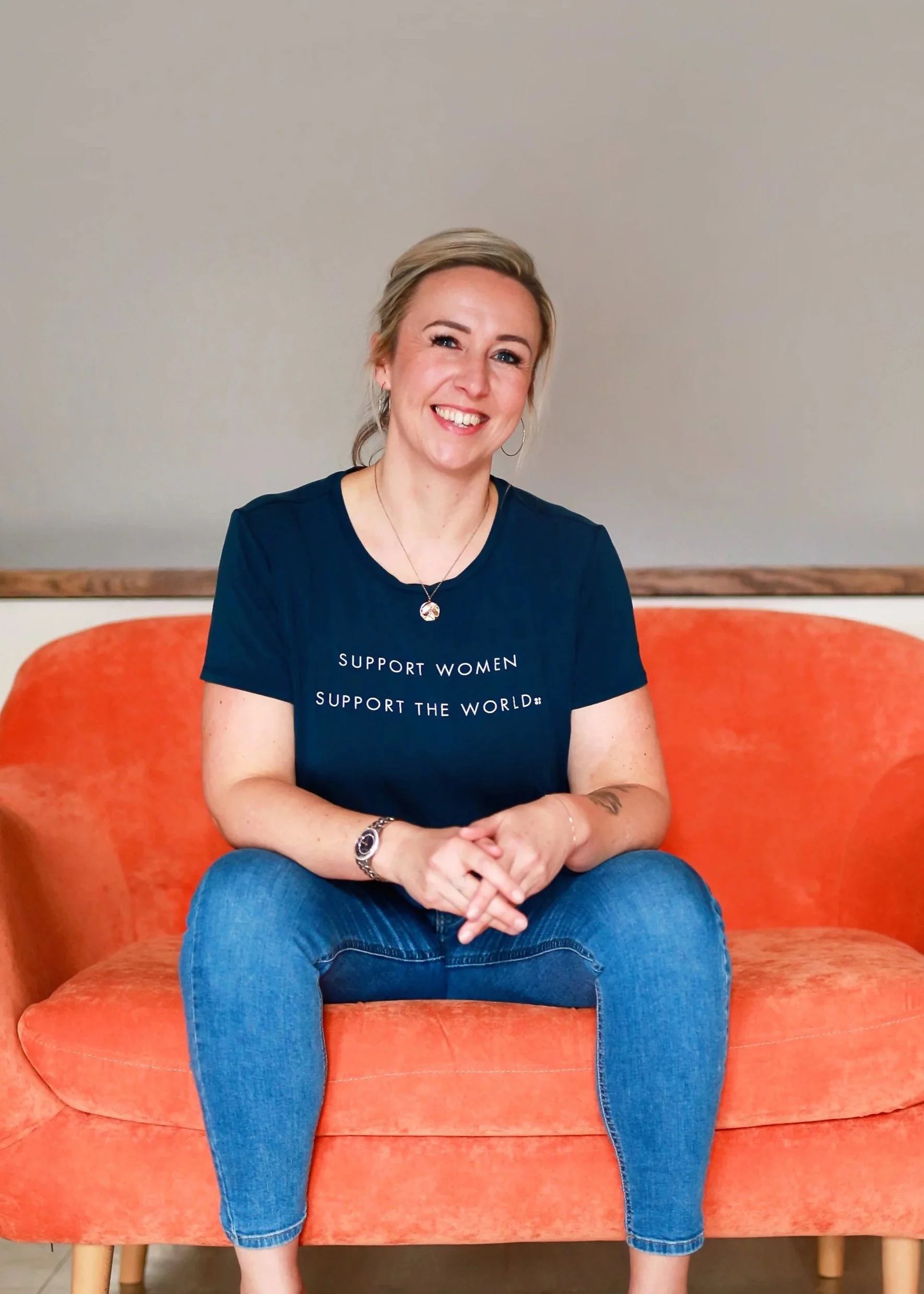 A smiling woman with blonde hair and blue eyes sitting on an orange couch against a plain beige wall, wearing a navy blue t-shirt with the words 'Support Women, Support the World' and blue jeans.