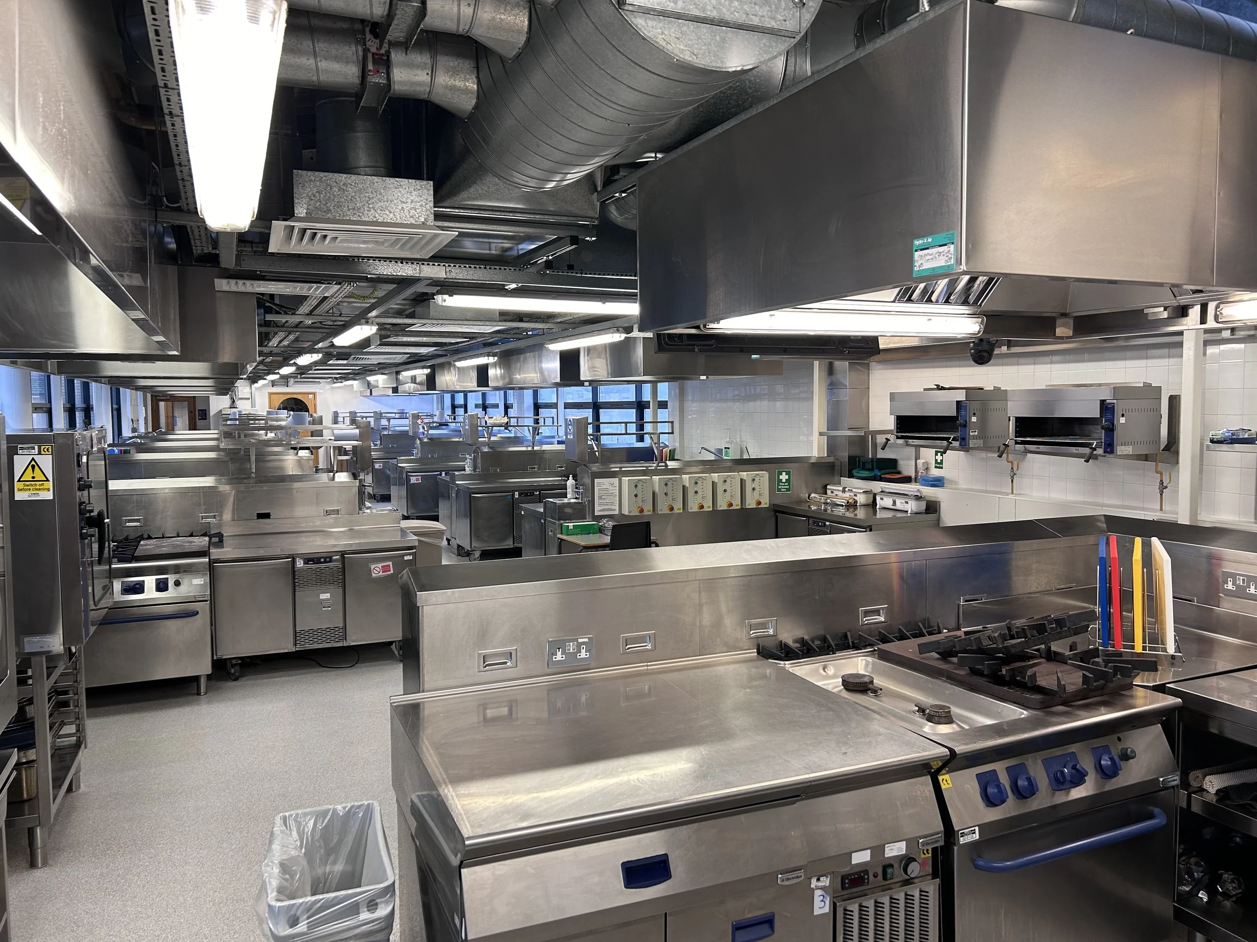OKN1's large professional commercial kitchen with stainless steel appliances, cooking stations, ovens, and cooking tools, illuminated by overhead lighting and large windows.