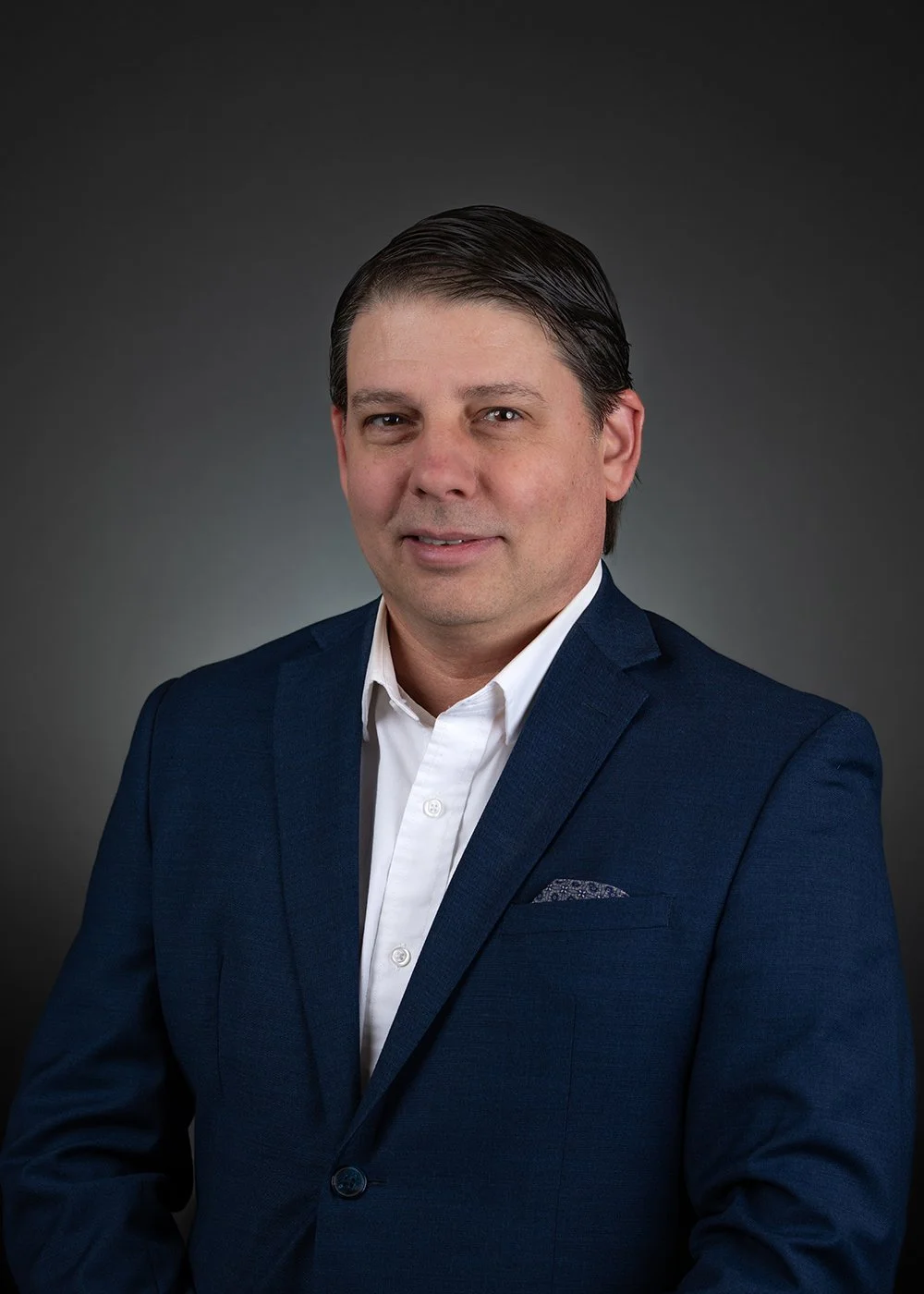Man in a suit with a blue tie smiling against a dark background.