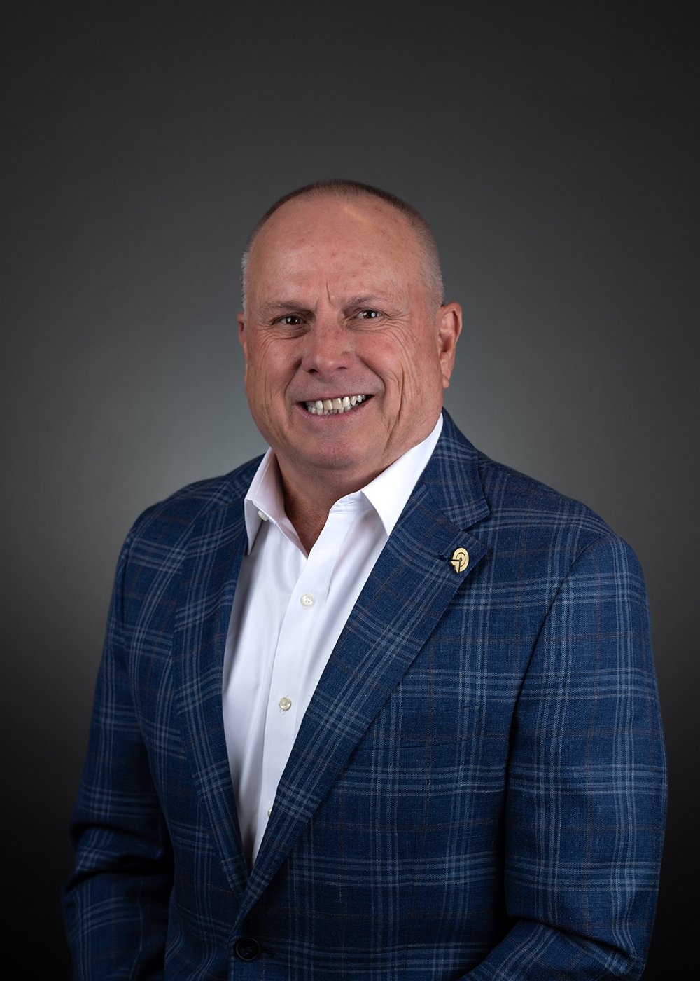 Smiling man in a suit with a light blue shirt and yellow tie, against a gray background.