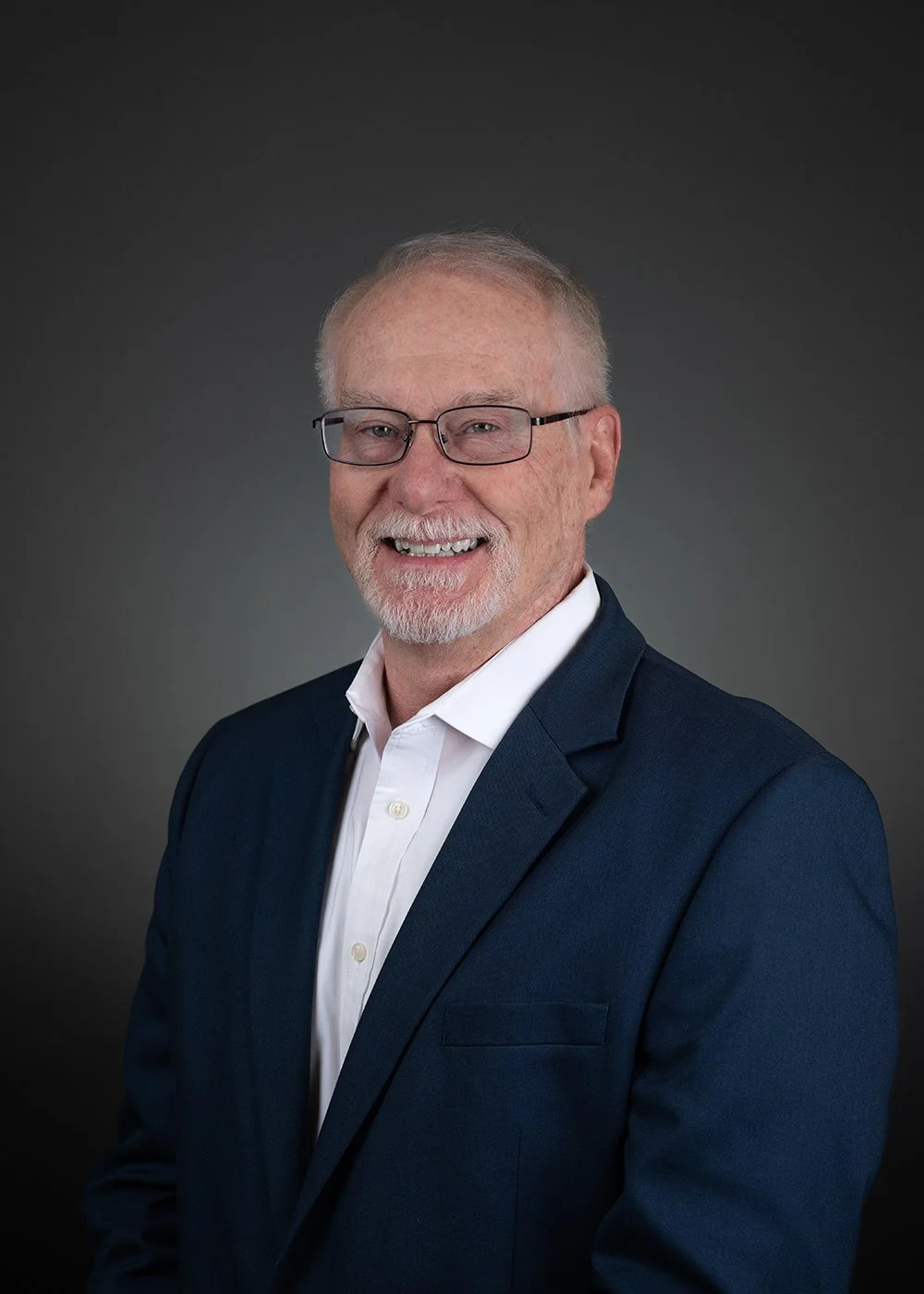 Elderly man in a suit with red tie, smiling, against a dark background.