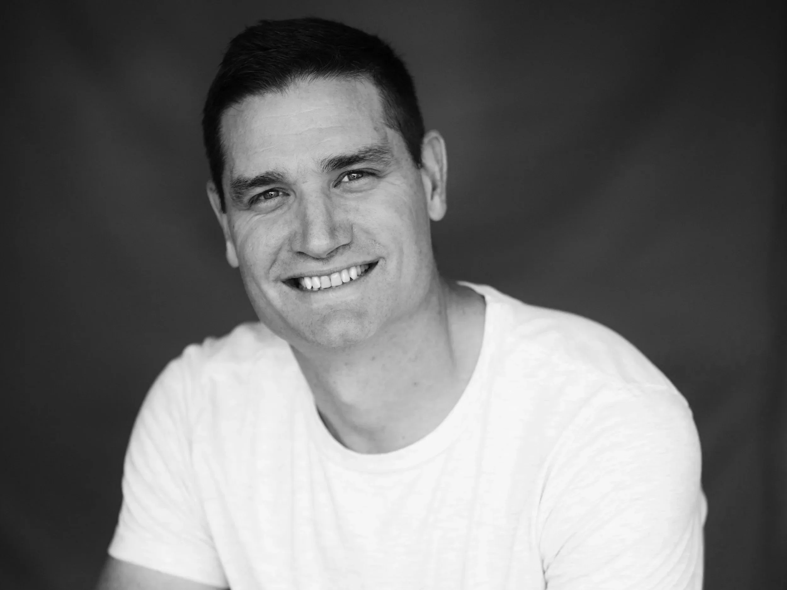 Black and white portrait of a smiling man with short hair, wearing a light-colored shirt, against a dark background.