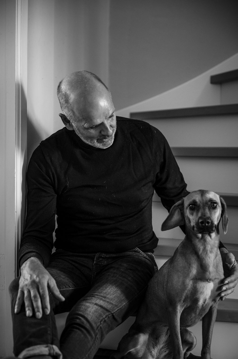 Ein Mann sitzt auf einer Treppe neben einem Hund. Der Mann trägt ein schwarzes Oberteil und Jeans. Das Bild ist in Schwarz-Weiß.