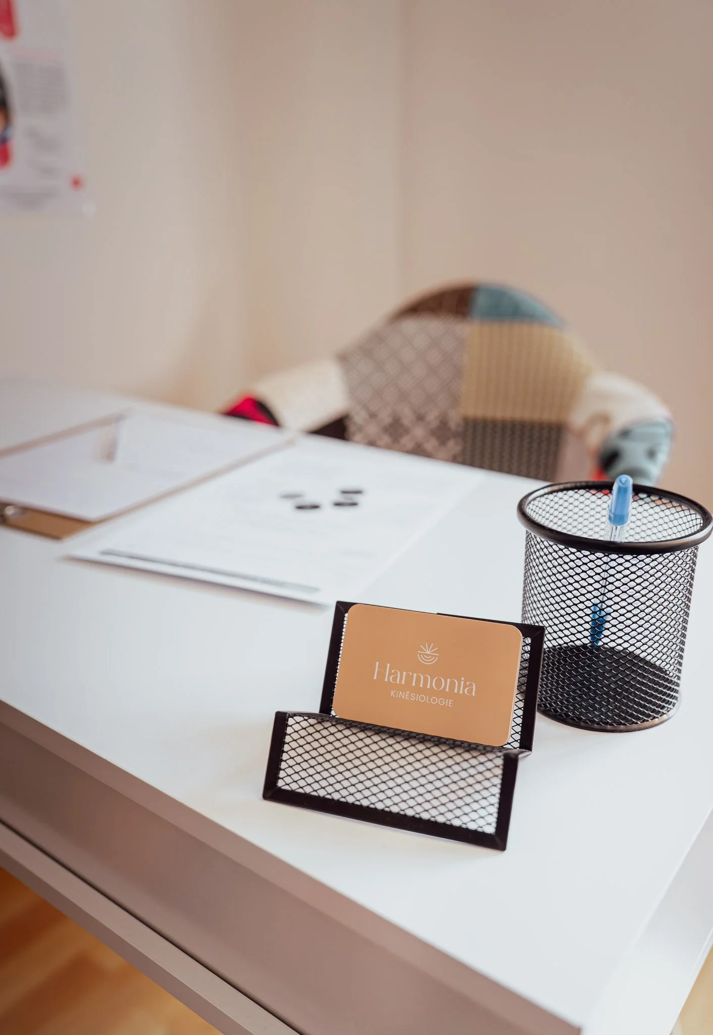 Un bureau blanc avec des documents, un pot à crayon, un porte carte avec une carte de visite "harmonia kinesiologie", un fauteuil coloré