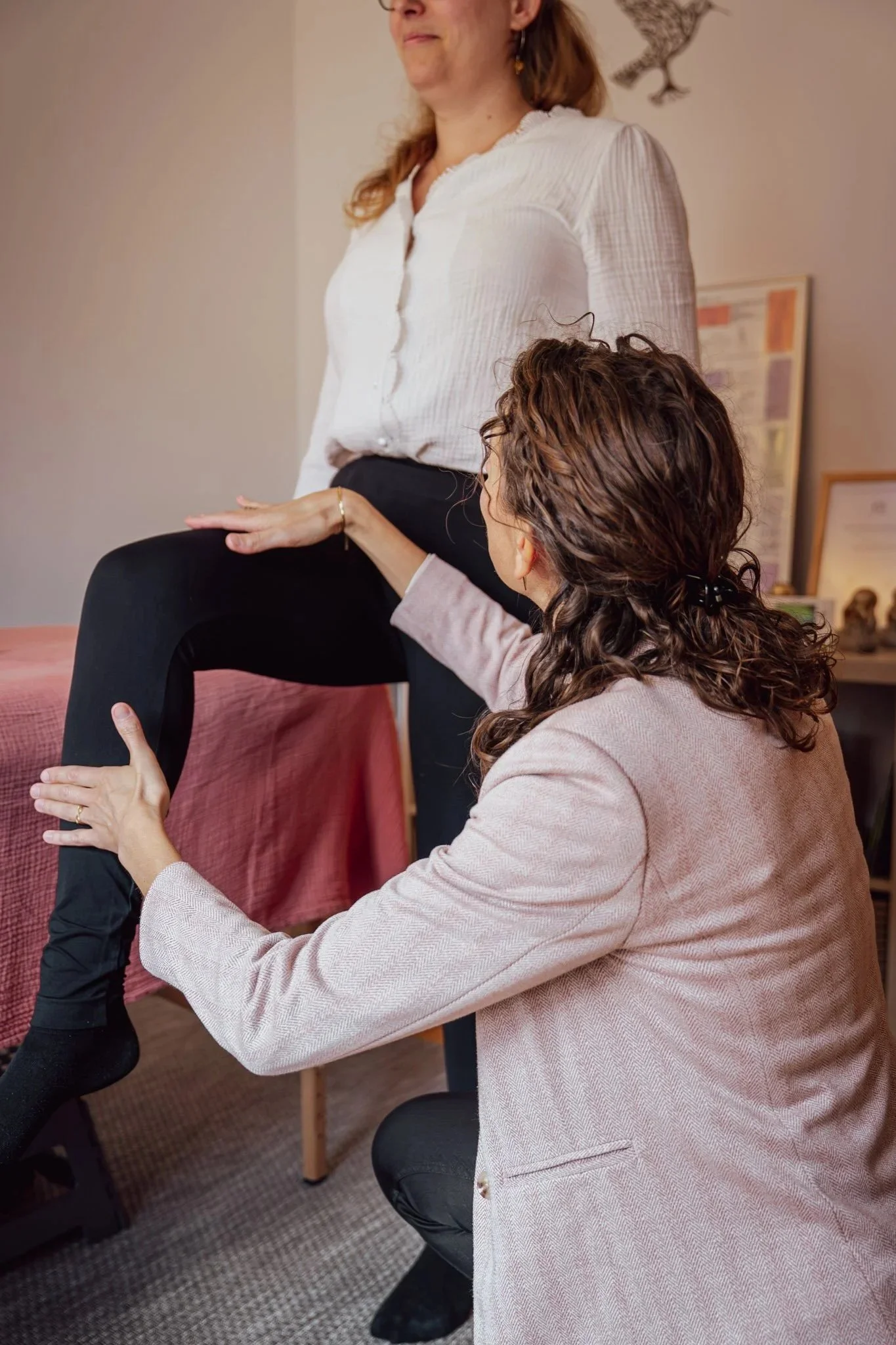 Une femme debout avec la jambe levée reçoit une séance de kinésiologie, l'autre femme accroupie la regarde et réalise un test musculaire sur la jambe.
