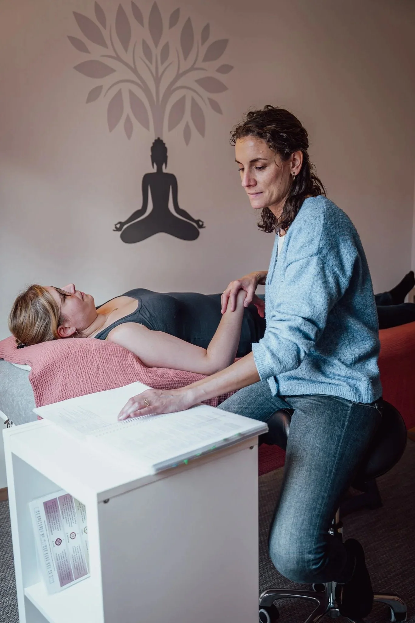 Une femme effectue un test musculaire de kinésiologie, tenant doucement le poignet d'une autre personne personne allongée sur la table de consultation. La femme, assise sur un tabouret, regarde des documents de kinésiologie posés sur un meuble blanc.
