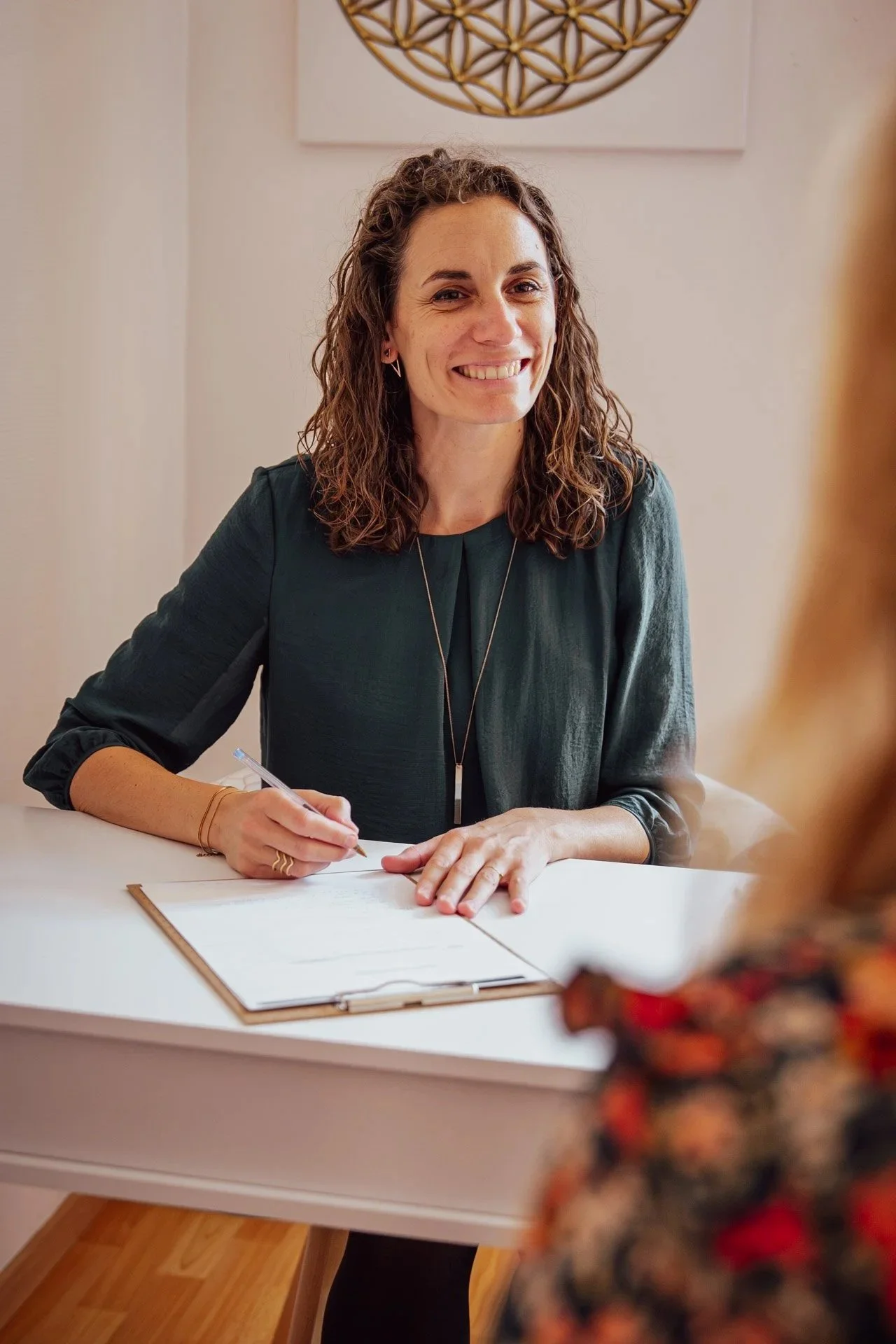 Une femme souriante en séance de kinésiologie, assise dans un fauteuil devant un bureau, parlant à une personne avec des cheveux blonds et un vêtement coloré, dans une pièce bien éclairée.