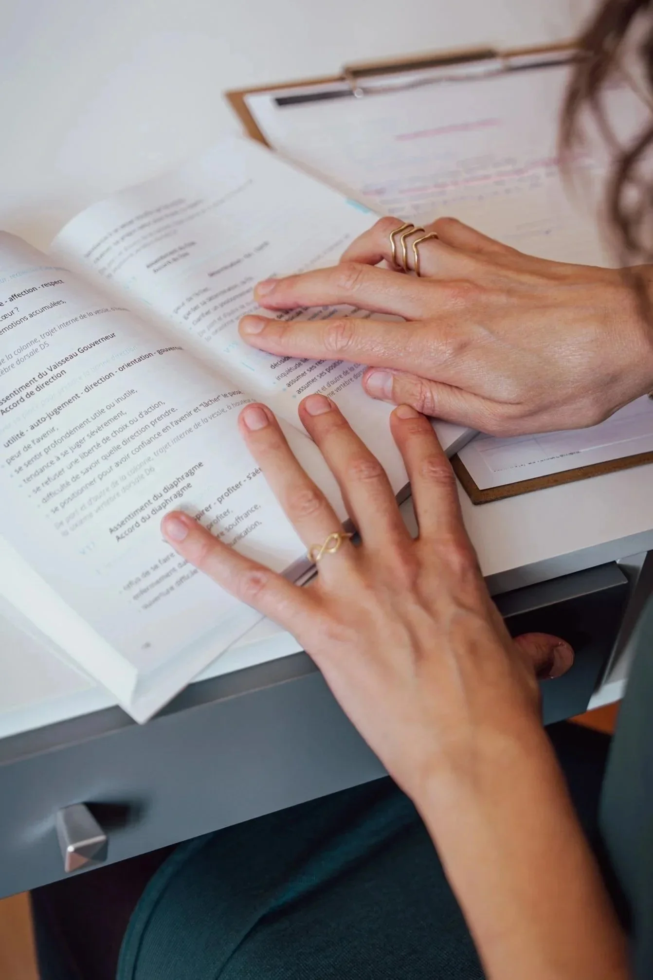 Sur un bureau, un livre sur les méridiens d'acupuncture est ouvert et une femme pose ses mains pour lire