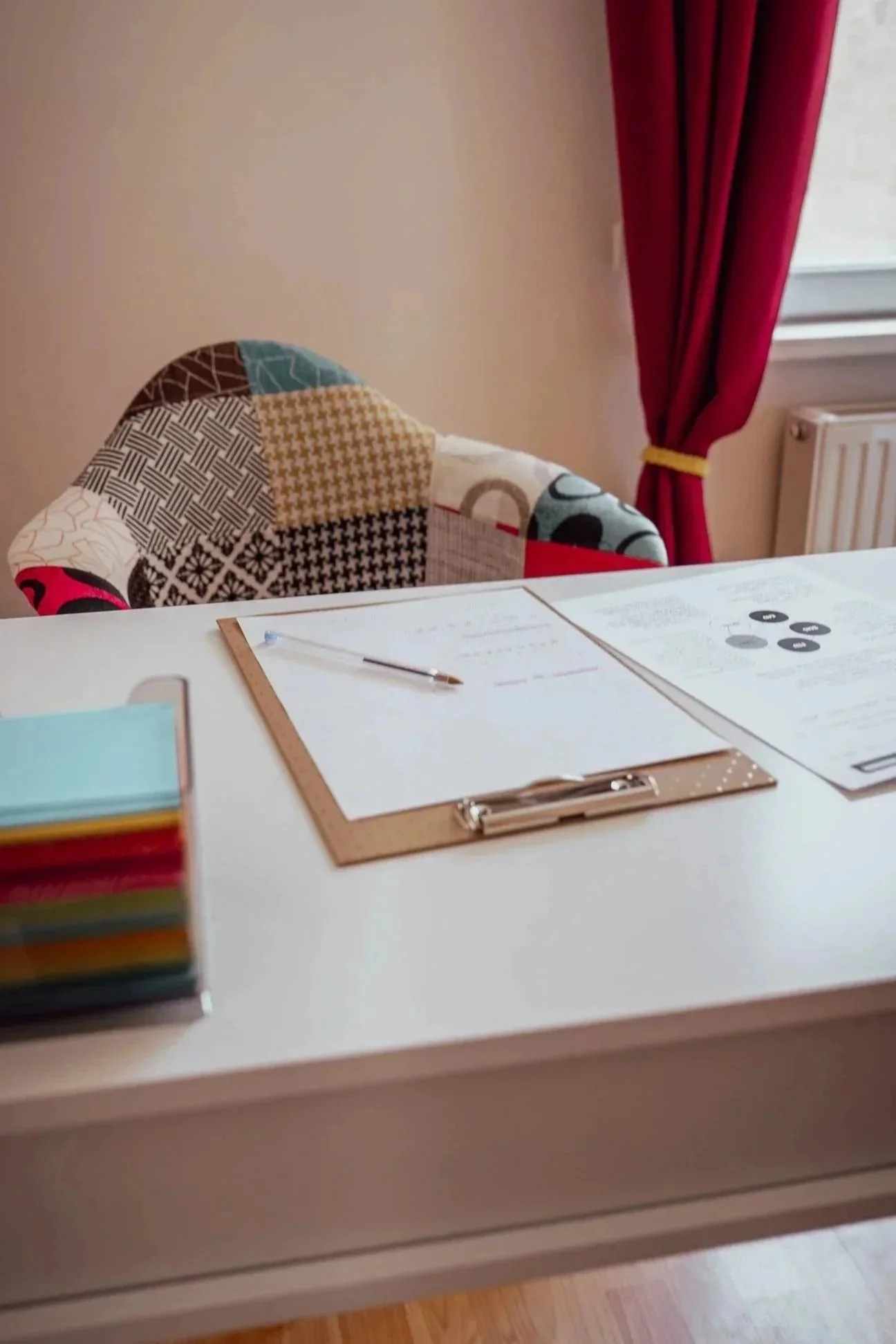 Un blanc bureau avec un bloc note coloré, des feuilles et un stylo et une fauteuil coloré avec  un rideau rouge dans une pièce lumineuse