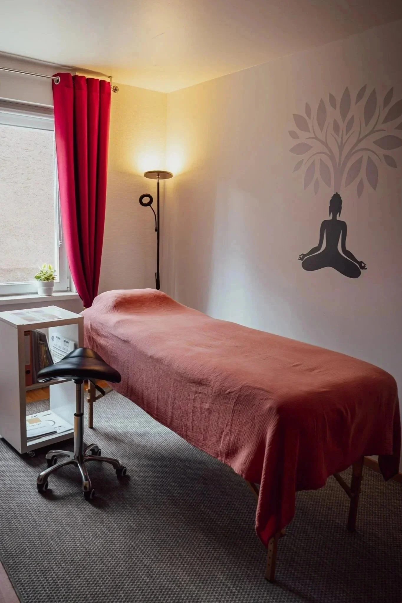 Une salle de consultation avec une table recouverte d'une housse orange, un meuble blanc, un tabouret, une lampe avec des rideaux rouges et un pais gris. Sur le mur, un dessin d'un arbre avec Bouddha.
