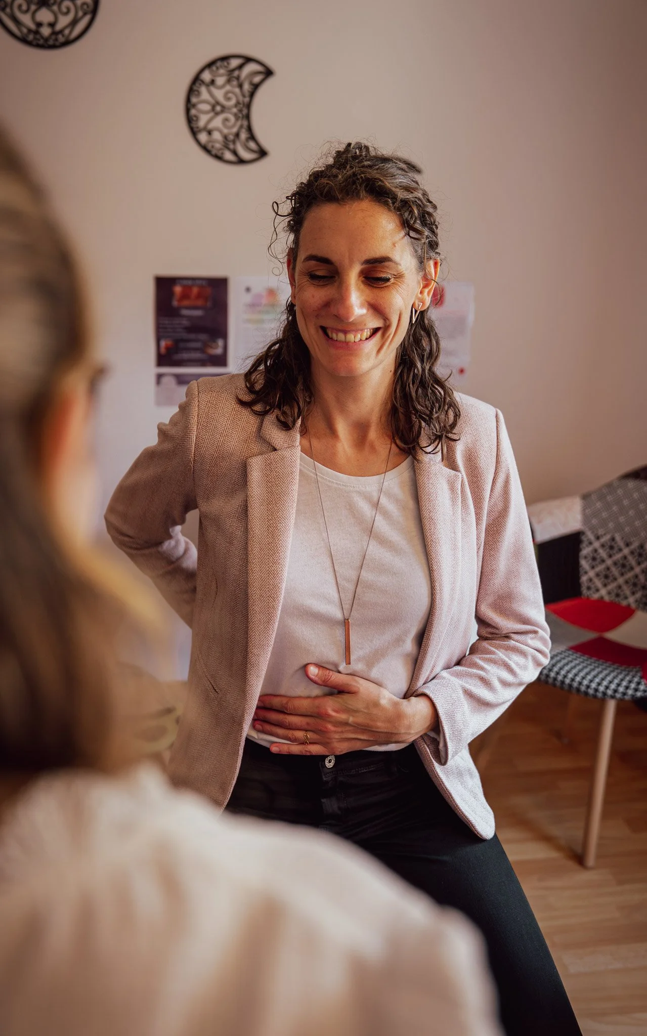 Femme assise avec un e main posée sur le ventre et l'autre main dans son dos. Face à elle une autre femme de dos avec des cheveux blonds. Une chaise colorée et des affiches au mur.