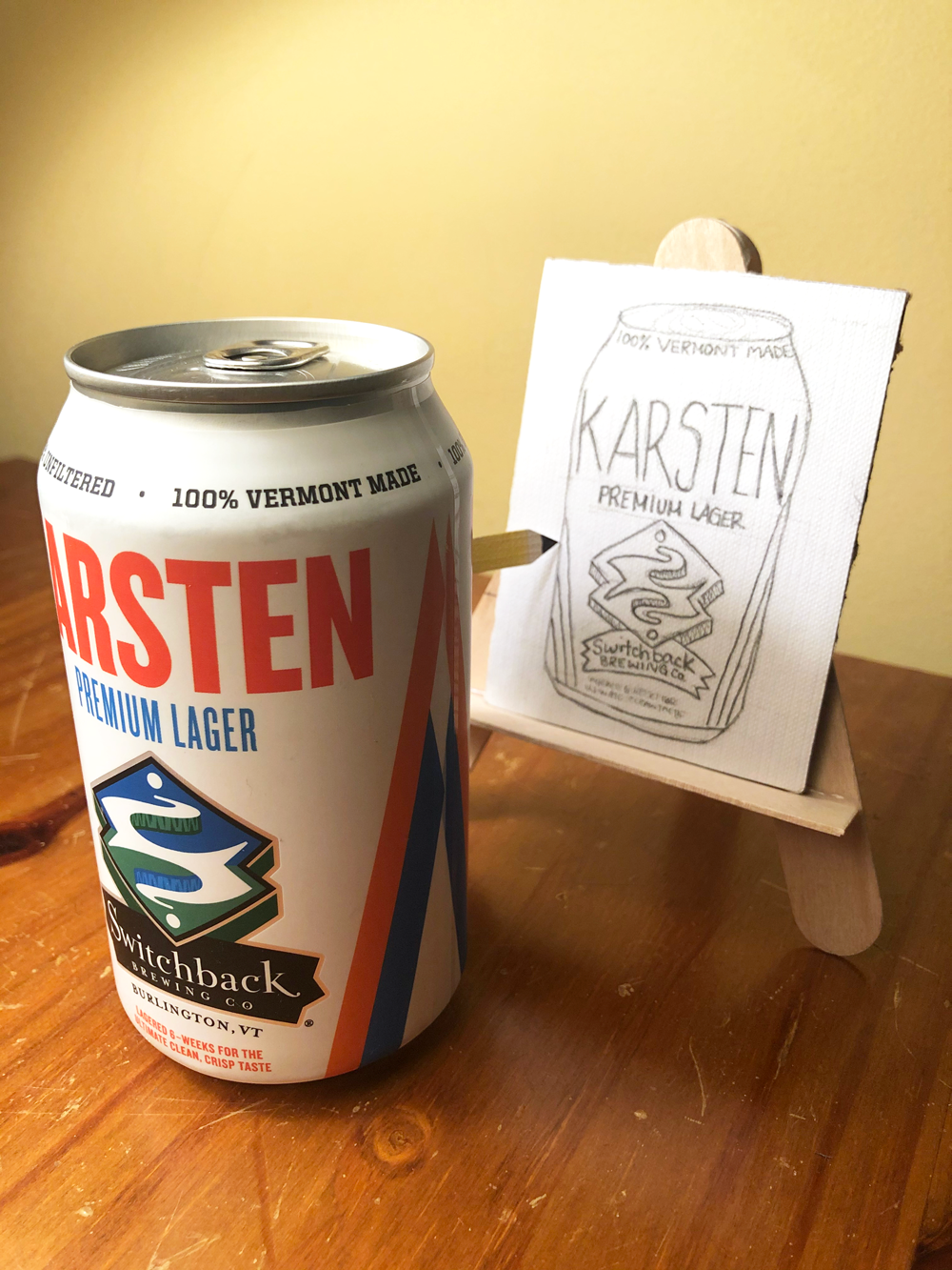 A can of Karsten Premium Lager by Switchback Brewing Co. next to a small easel displaying a pencil sketch of the can on a wooden surface.