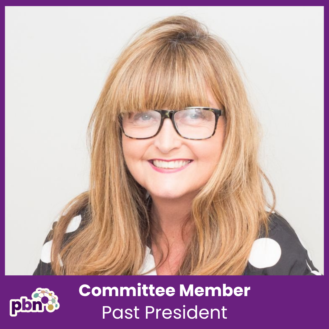 A woman with long red hair, glasses, and a black and white polka dot blouse sitting at a desk, smiling at the camera.