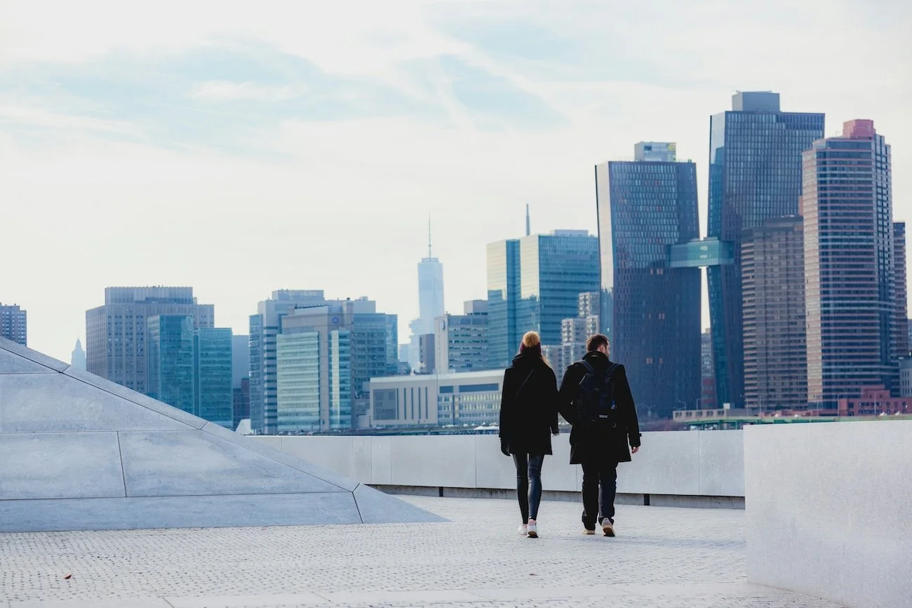Two people walking on a rooftop with a city skyline in the background.