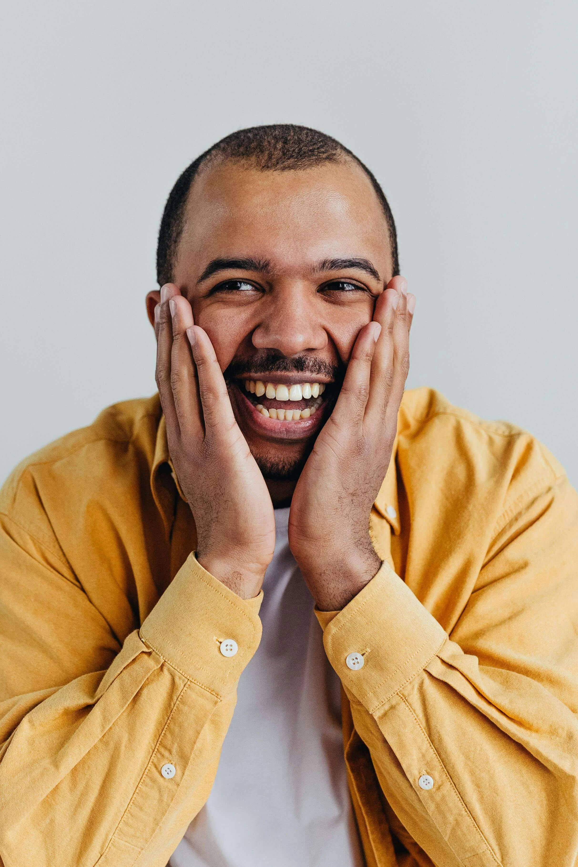 Man in yellow shirt smiling with hands on face against neutral background. Build sustainable systems that work with your brain through twice exceptional therapy in NYC.