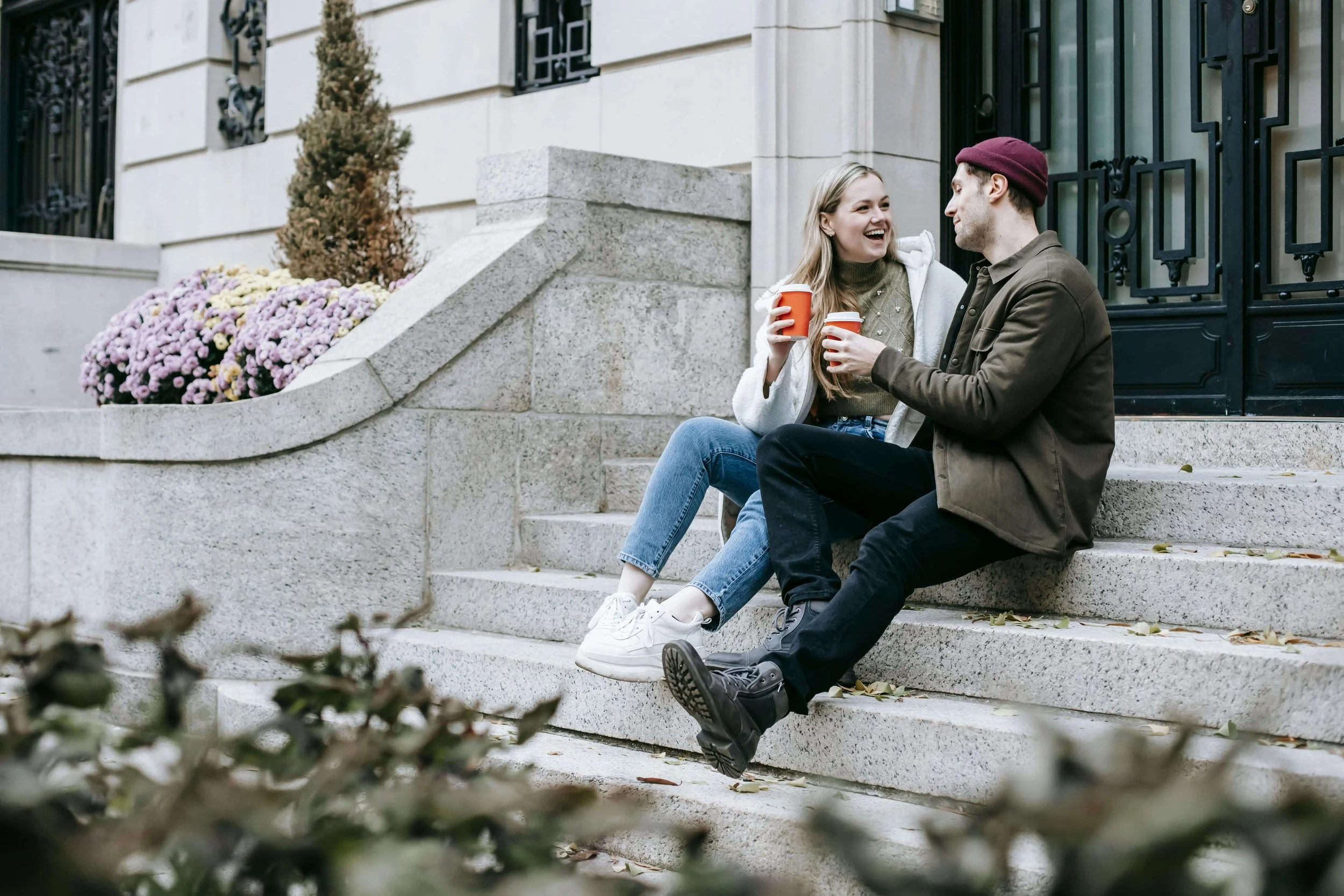 A smiling couple sitting on stone steps outdoors, holding coffee cups and talking. Rediscover joy and connection in your relationship with the guidance of a couples therapist in NYC.