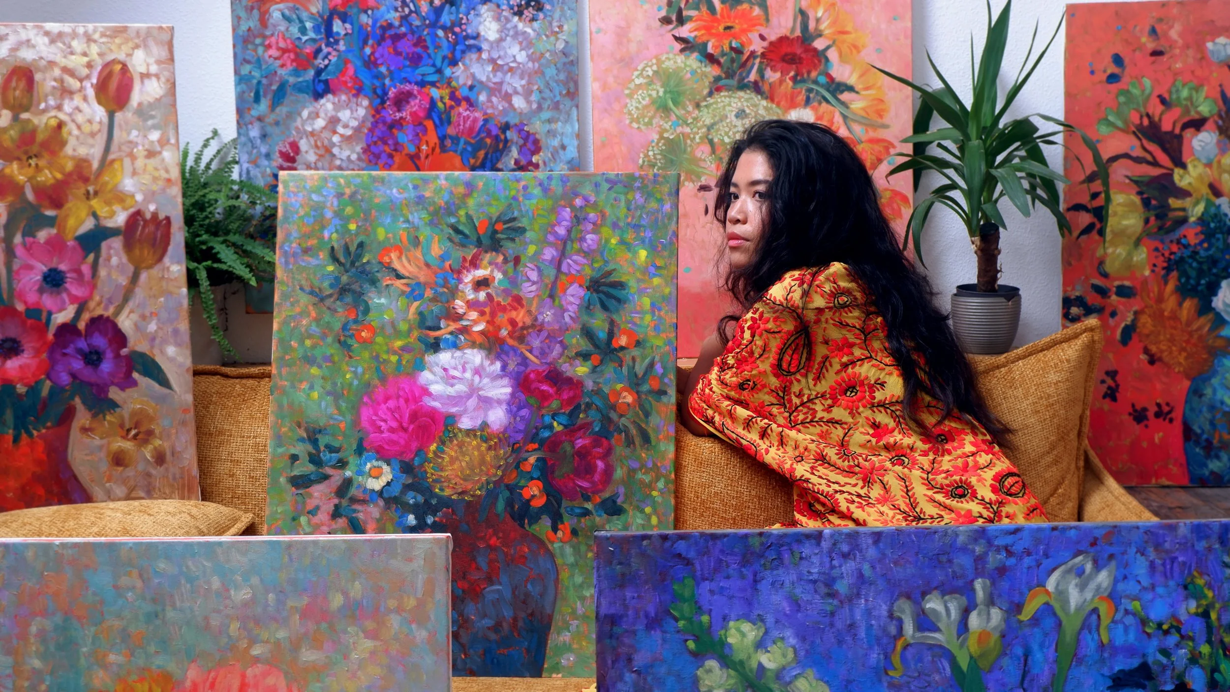 A woman with long black curly hair wearing a yellow dress with red floral patterns sitting on a tan couch surrounded by colorful floral paintings in an art studio or gallery.
