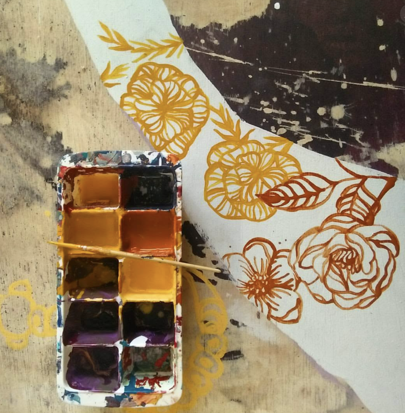 A watercolor palette with six colors including shades of yellow, purple, and brown, set next to a painting of flowers and leaves in yellow and brown on fabric with a splattered dark background.
