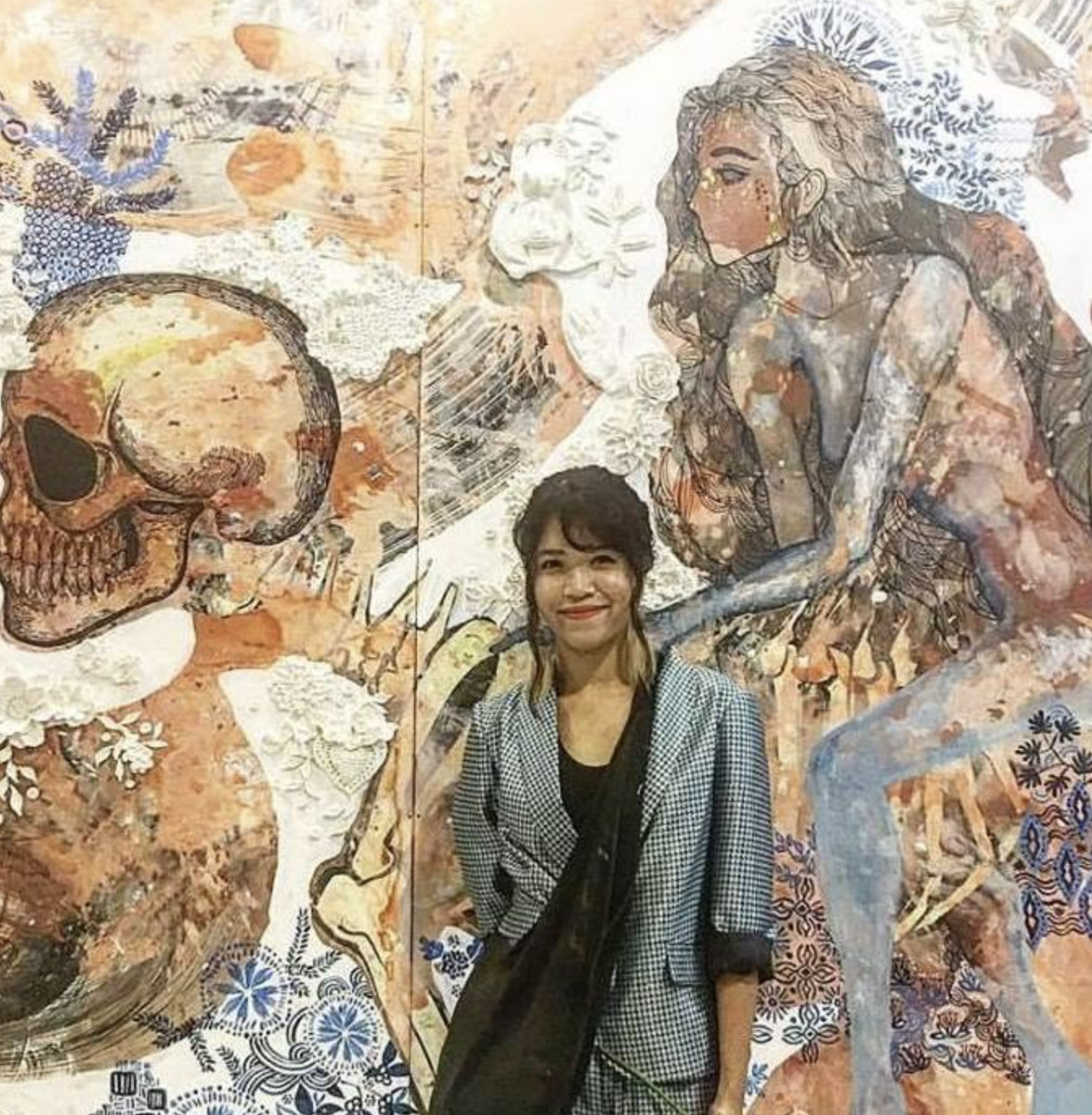 A woman standing in front of a mural featuring a skull, a woman, and abstract figures.