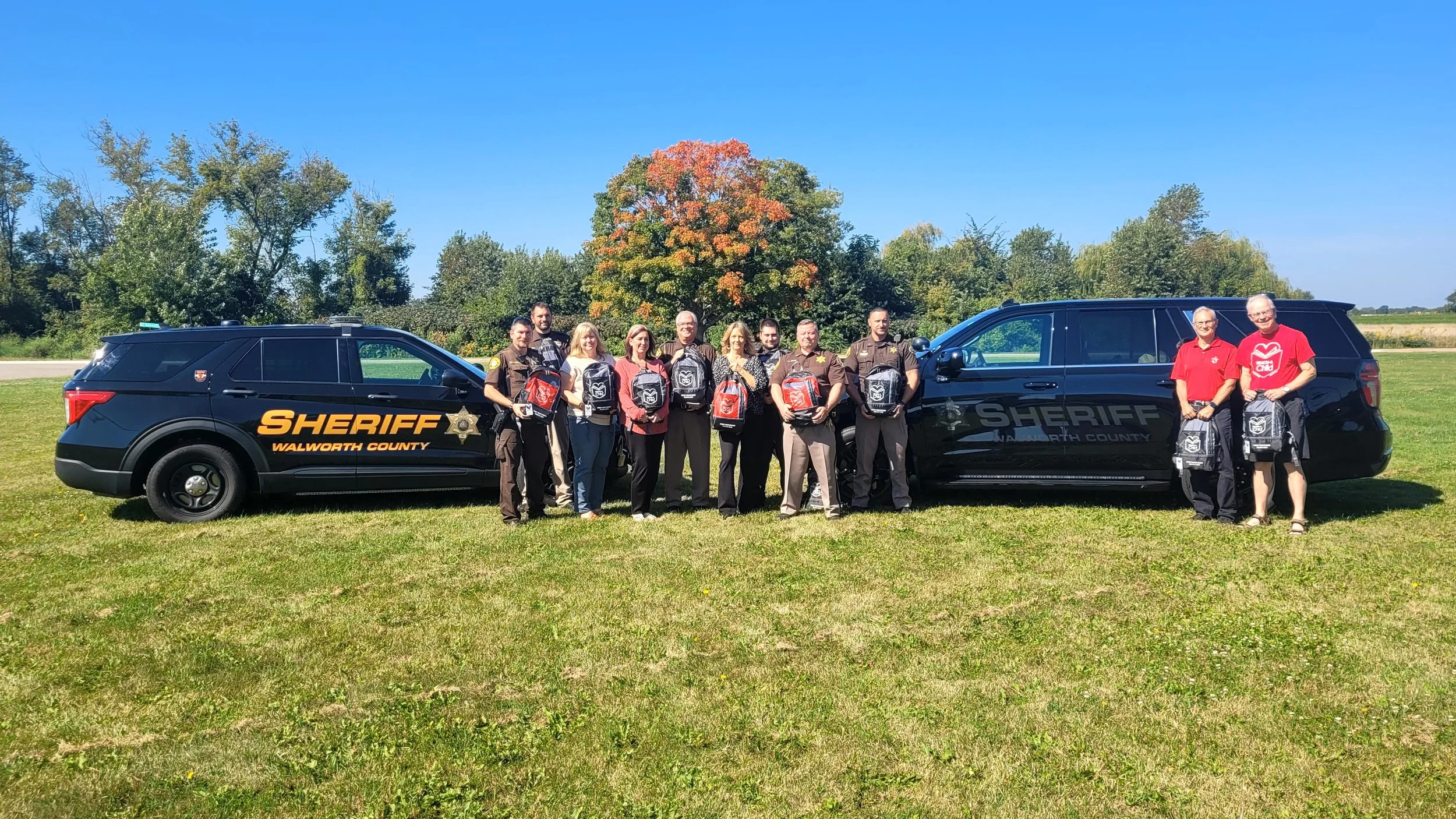 REACH-A-CHILD with REACH BAGs with the Walworth County Sherif's Office in front of police cars on the grass