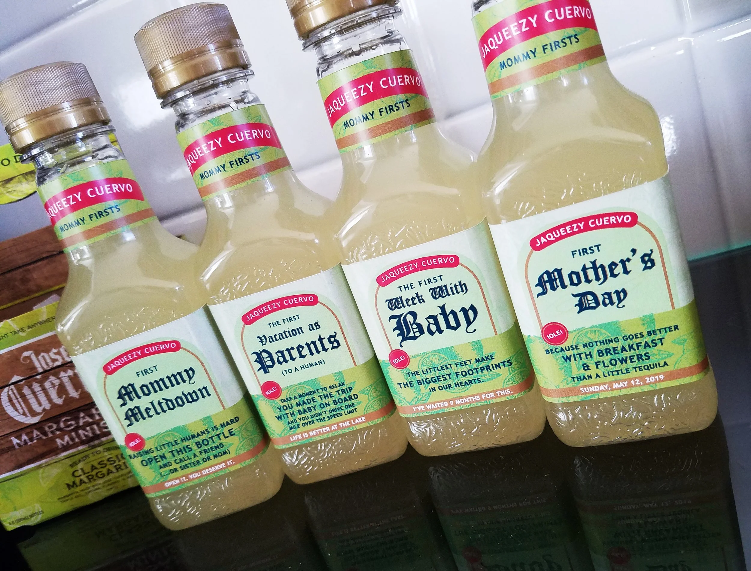 Five bottles  tequila mix, each with humorous labels related to parenting, including 'First Mommy Meltdown', 'First Vacation as Parents', 'First Week with Baby', and 'First Mother's Day', displayed on a reflective surface.