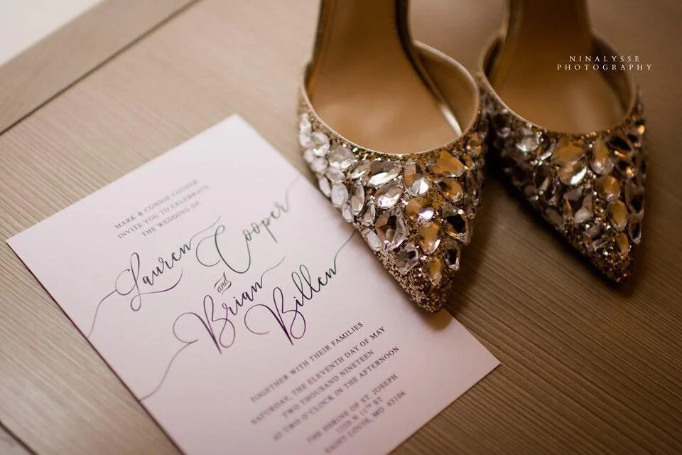 Close-up of sparkly high-heeled shoes next to a wedding invitation on a wooden surface.