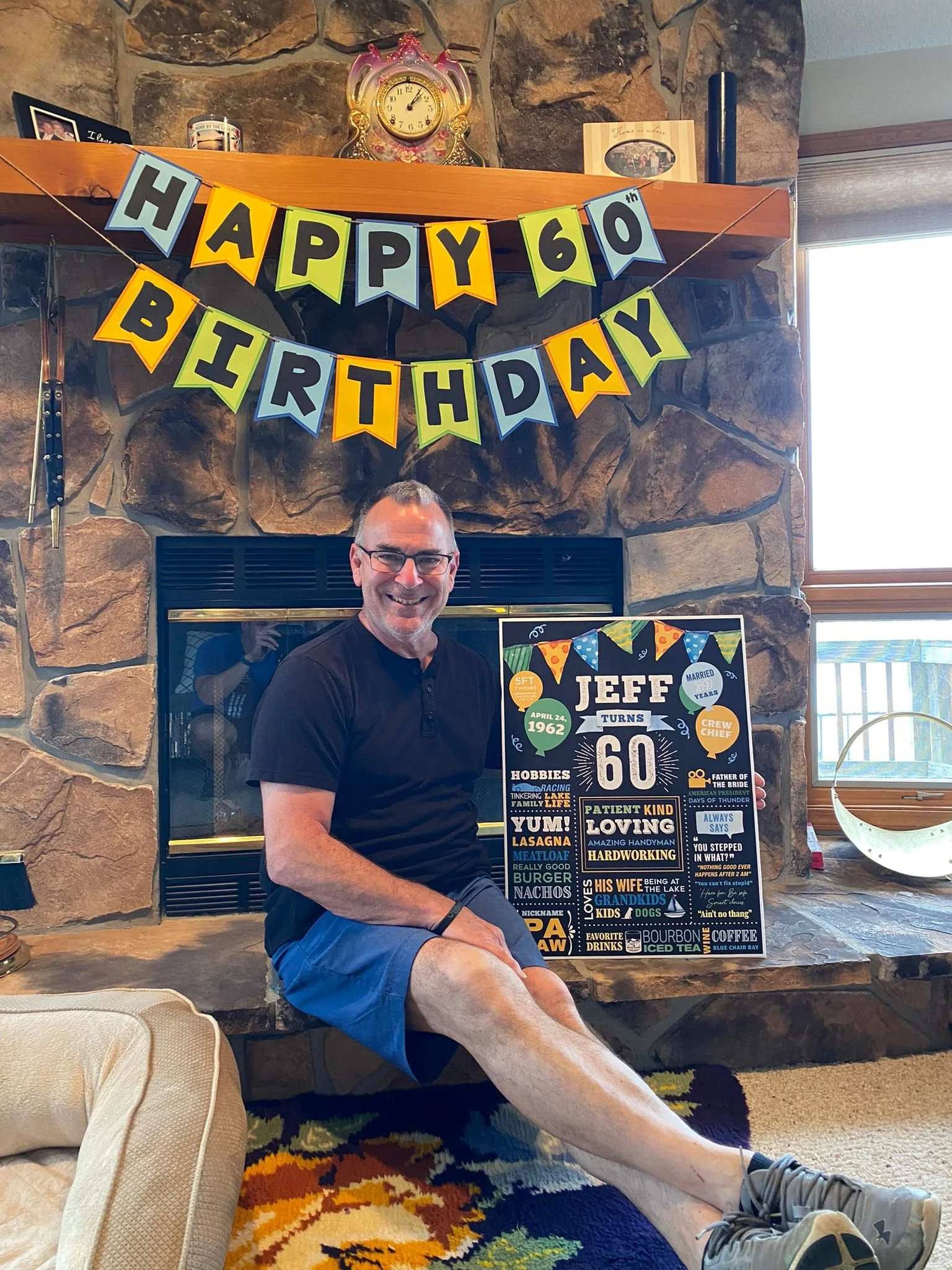 A man sitting in front of a fireplace with a "Happy 60th Birthday" banner. He is holding a poster with personal interests and information, such as hobbies and favorite foods. There's a rug on the floor and a clock on the mantel.
