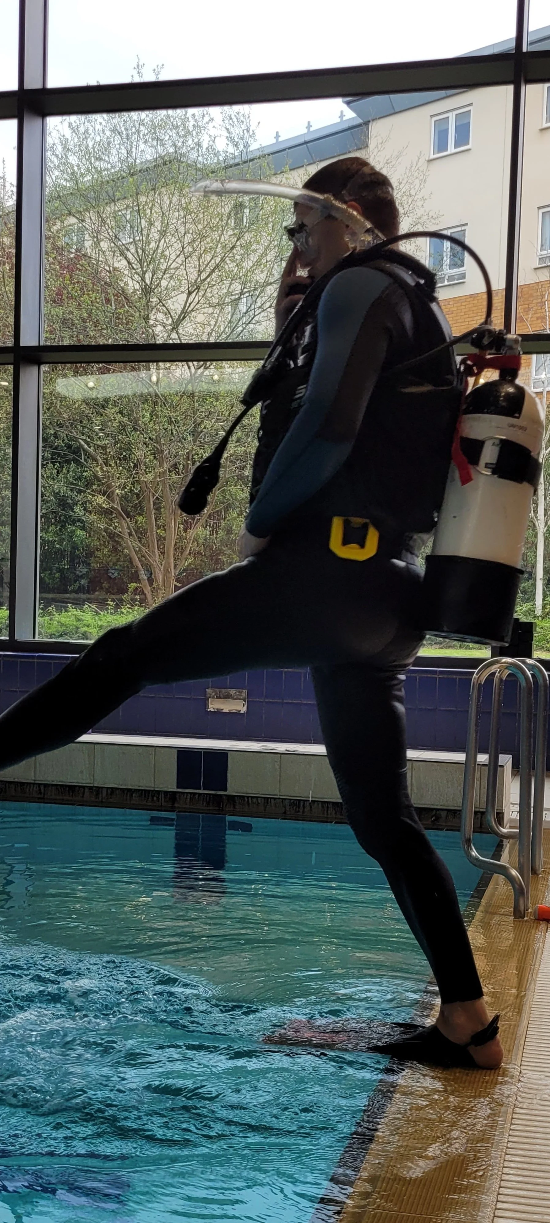 Person in wetsuit with scuba gear standing at the edge of an indoor swimming pool, preparing to dive in, with large window showing trees and residential buildings outside.