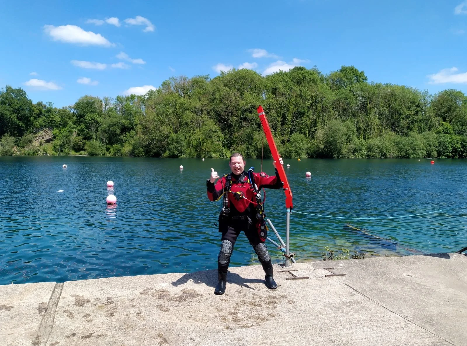 Man in scuba gear holding a flotation device near a lake with trees and a blue sky in the background.