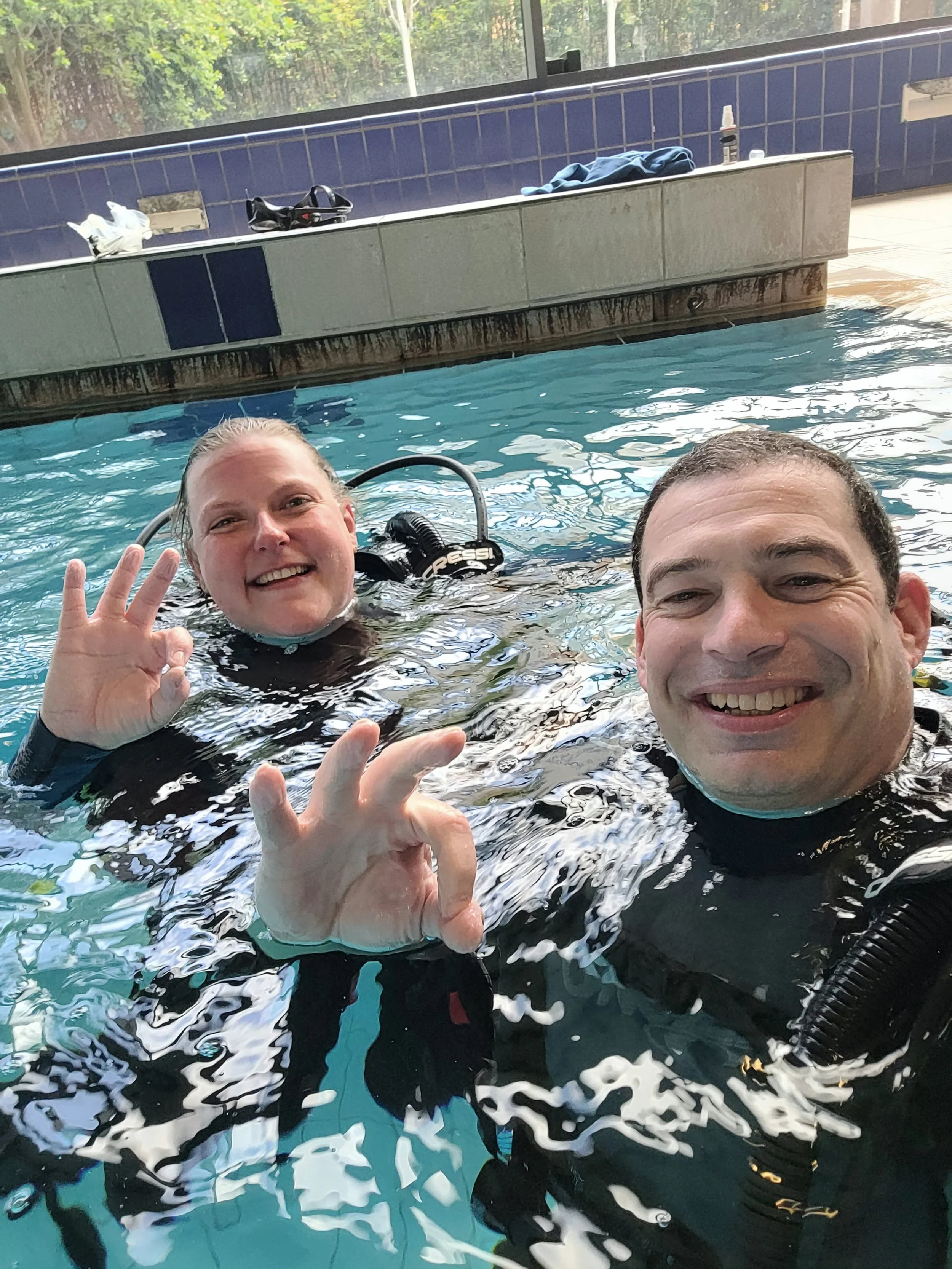 A man and a woman in a swimming pool, smiling and making the 'OK' sign with their hands, with items like towels and personal belongings on the poolside behind them.