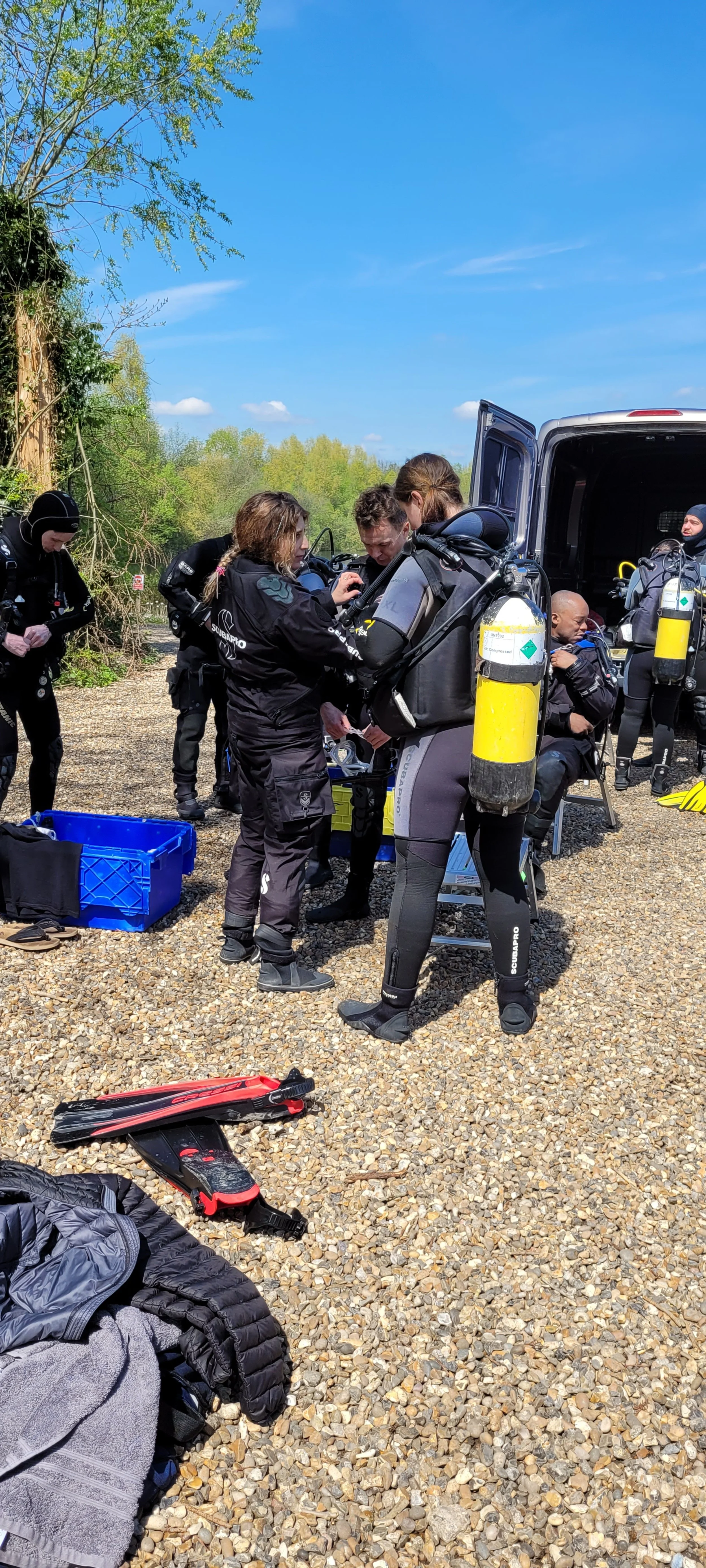 A group of scuba divers in wetsuits preparing gear outdoors on a gravel surface, with equipment, a blue storage bin, and a red fins paddle visible, near a van and a wooded area.