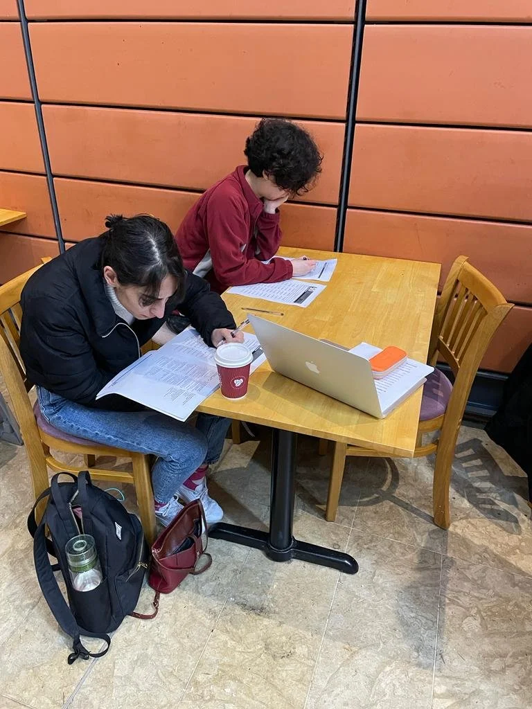 Two students sitting at a wooden table studying with open textbooks, a laptop, and a coffee cup in a room with orange paneled walls.