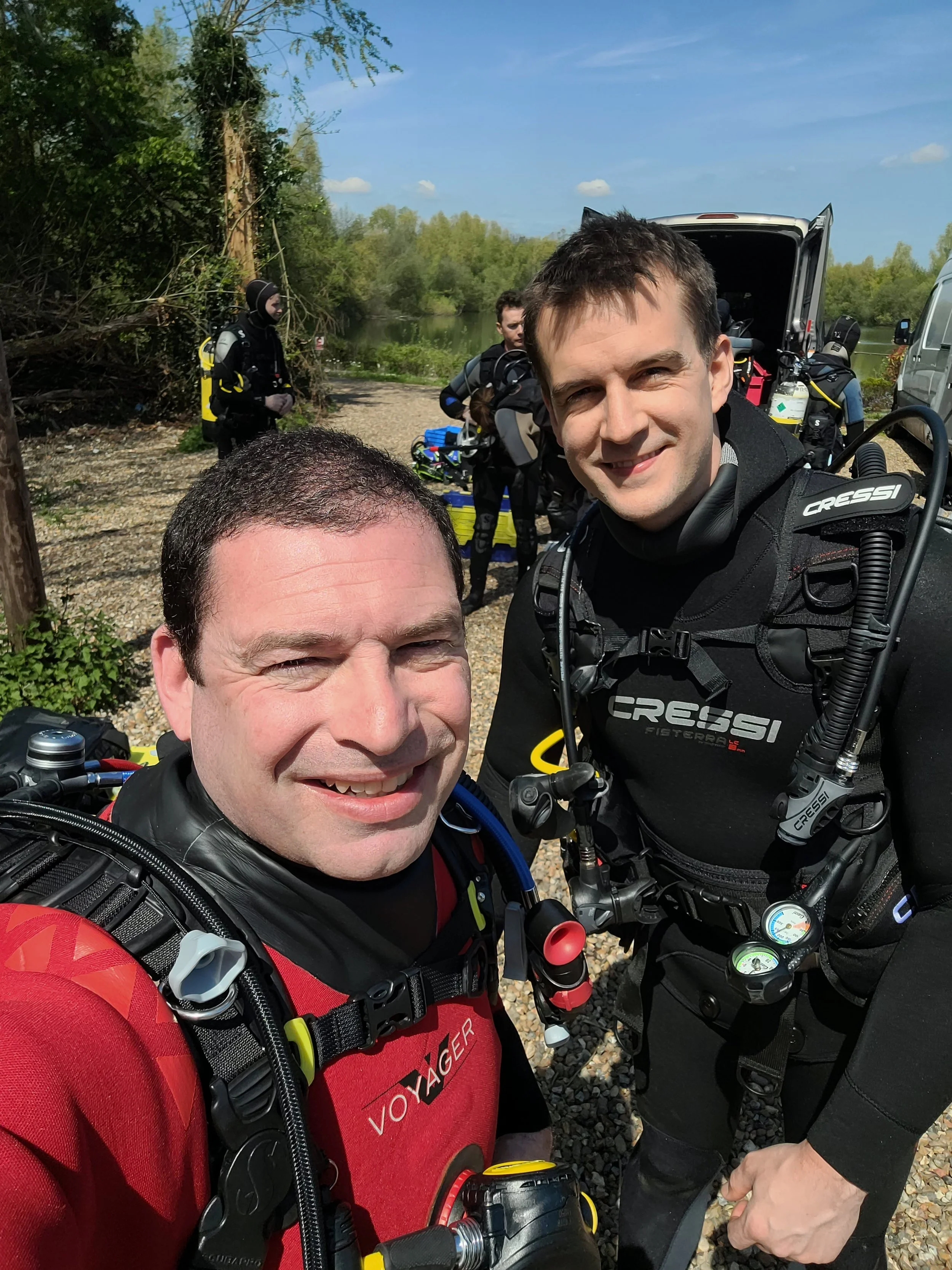 Two men in scuba gear taking a selfie outdoors near a body of water, with other divers preparing equipment in the background.