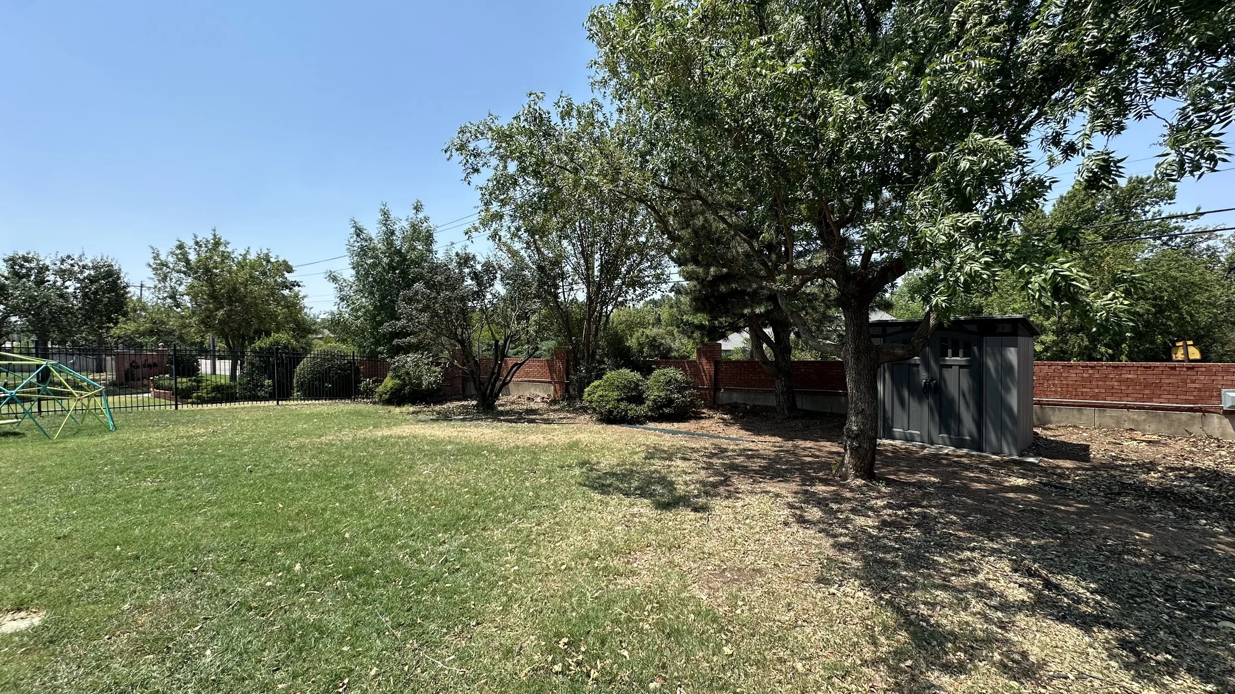 A backyard with green grass, a large tree, and a small shed, enclosed by a brick and metal fence, under a clear blue sky.