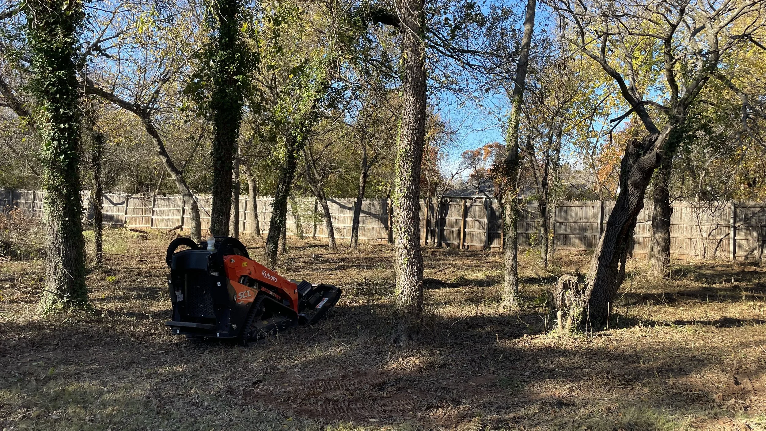 Backyard with a wooden fence, trees with some green leaves, and a small orange Kubota stump grinder machine on the ground.