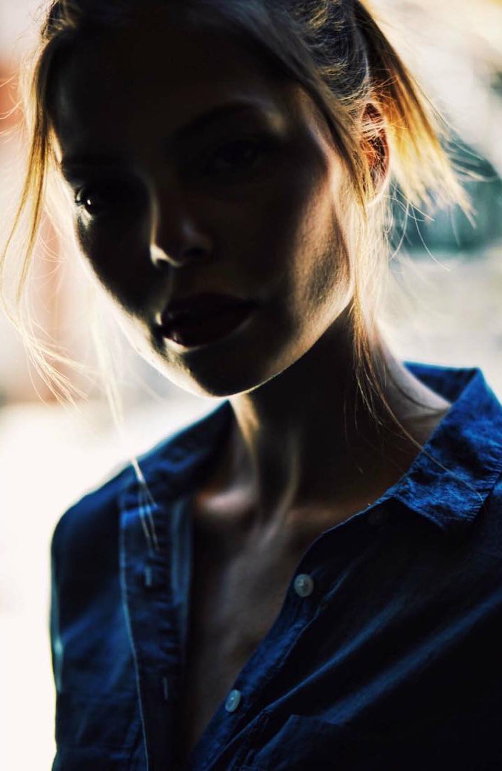 A woman with blonde hair and light skin, wearing a blue denim shirt, is in a backlit setting, casting her face in shadow.