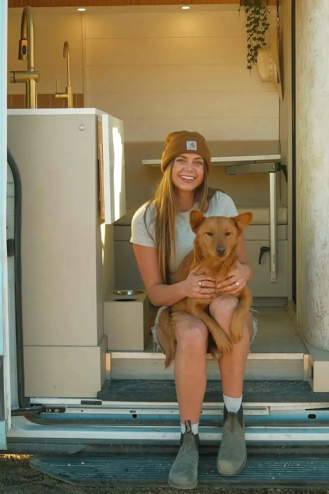 A woman sitting on the steps of a small camper or tiny house, holding a brown dog on her lap. She is smiling, wearing a gray t-shirt, gray shoes, a tan beanie, and white socks.