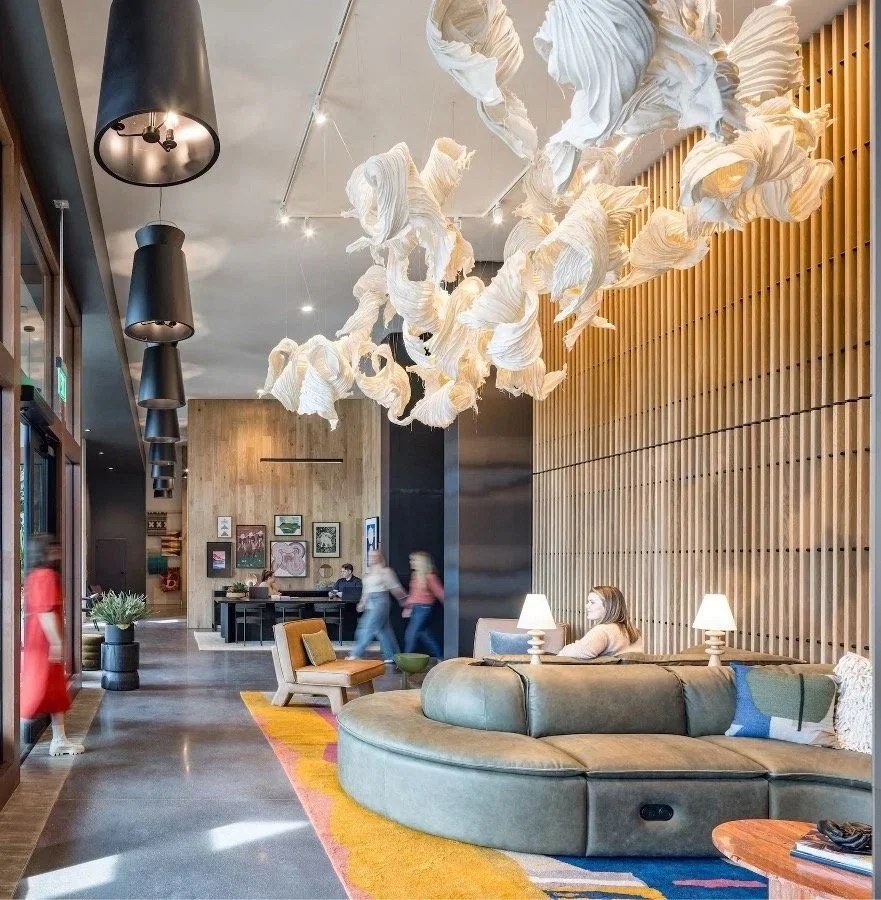 A modern hotel lobby with a large circular gray sofa, two side lamps, and a textured wood wall. A woman sits on the sofa, and abstract white fabric art hangs from the ceiling. Several people walk through the space.