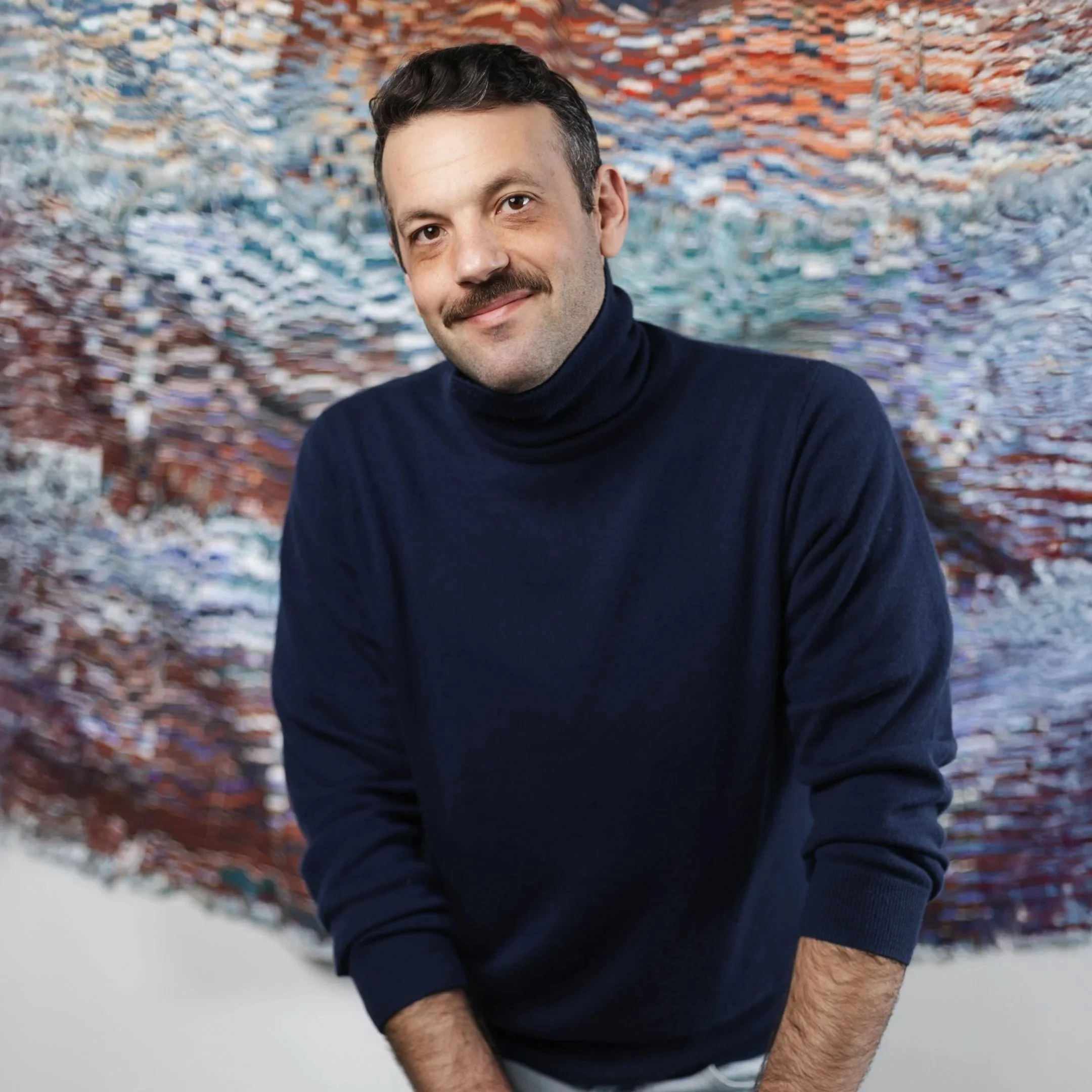 Portrait of a man with dark hair, a mustache, wearing a black turtleneck sweater, standing in front of an abstract, colorful background.