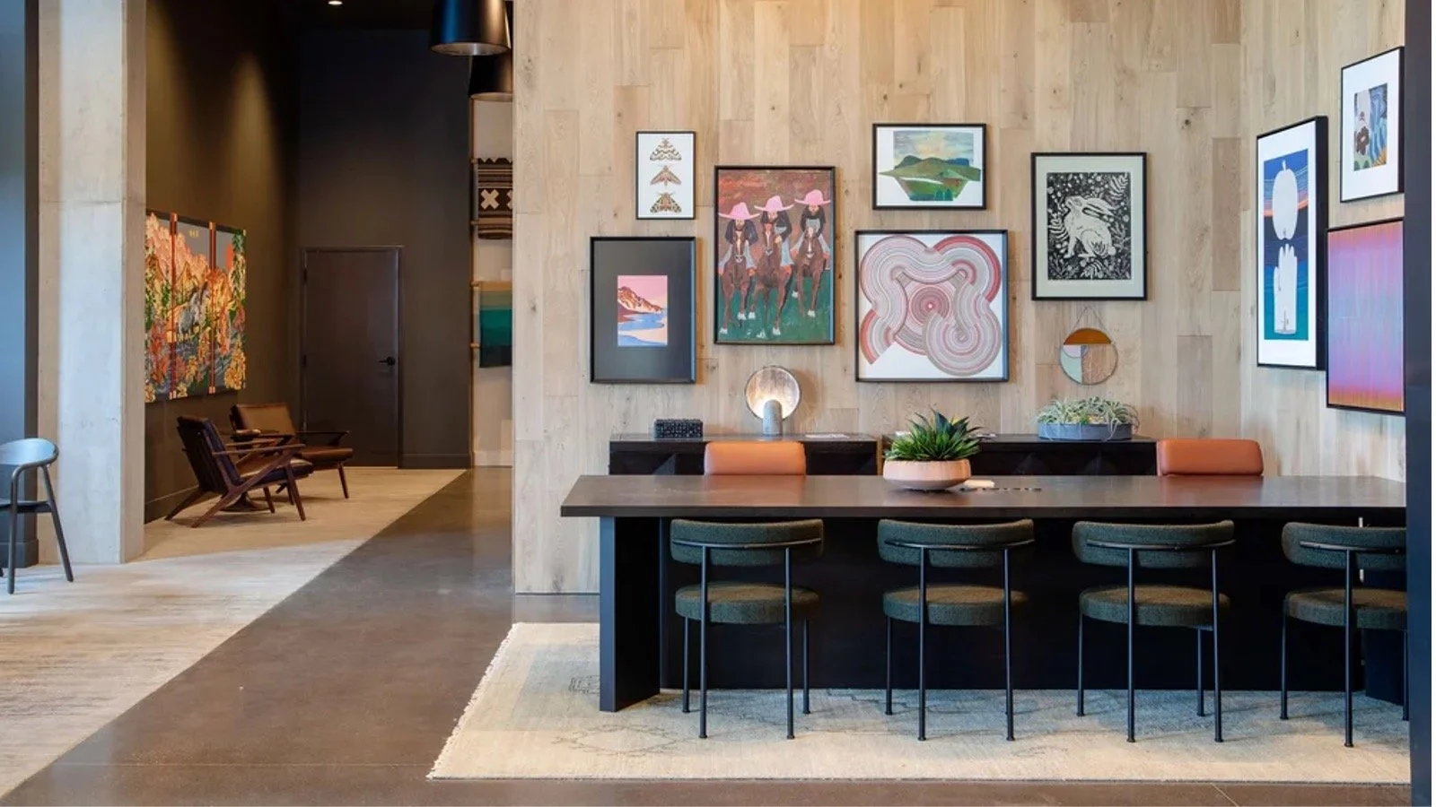 Interior of a modern art gallery or lounge area with a wooden wall adorned with framed artwork, a table with chairs, and a seating area with wooden chairs and artwork in the background.