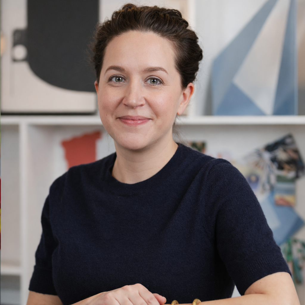 A woman with short brown hair and light skin smiling at the camera in an indoor setting with artwork and shelves in the background.