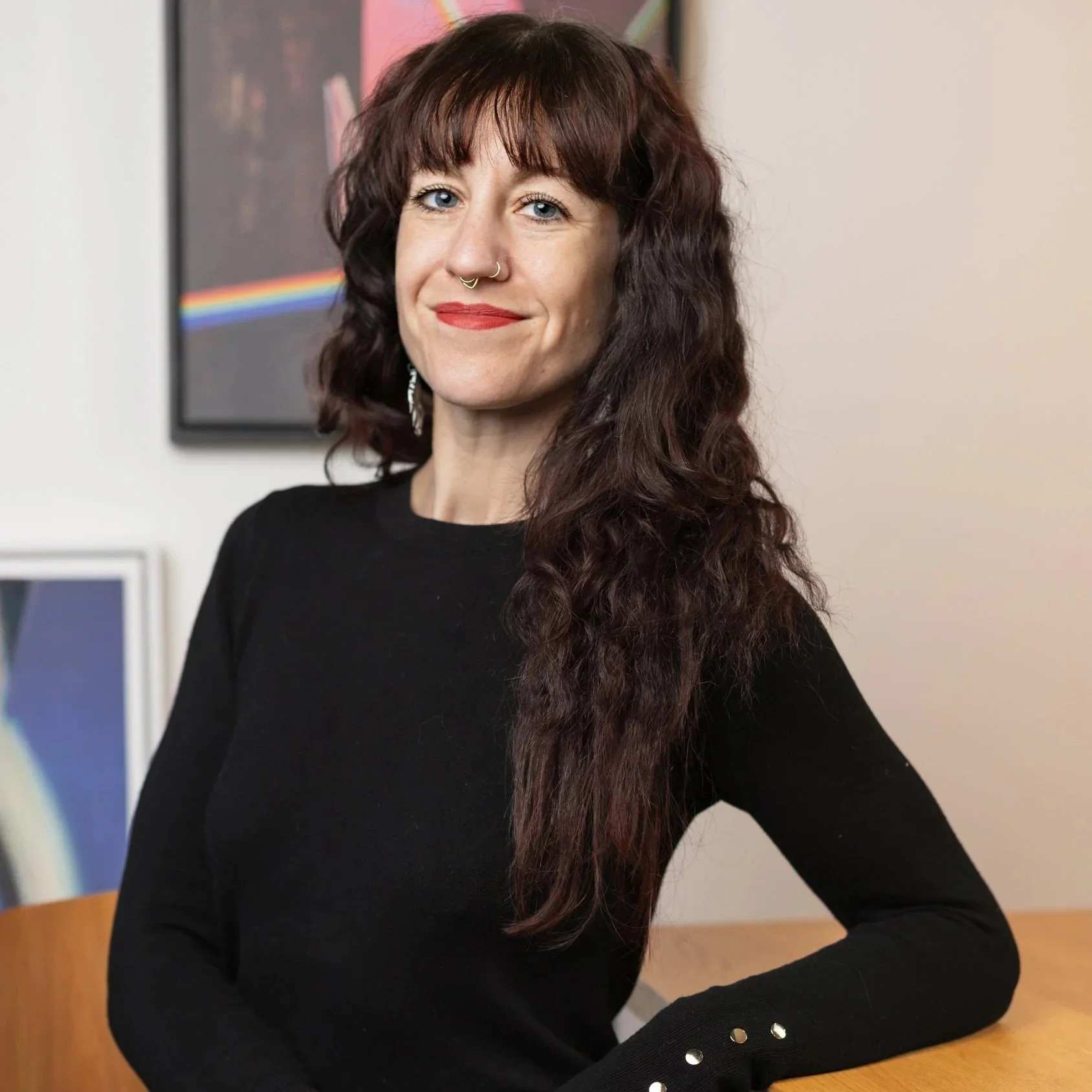 A woman with long curly brown hair, wearing a black top with metallic buttons on the sleeve, sitting at a wooden table with a neutral background and artwork on the wall.