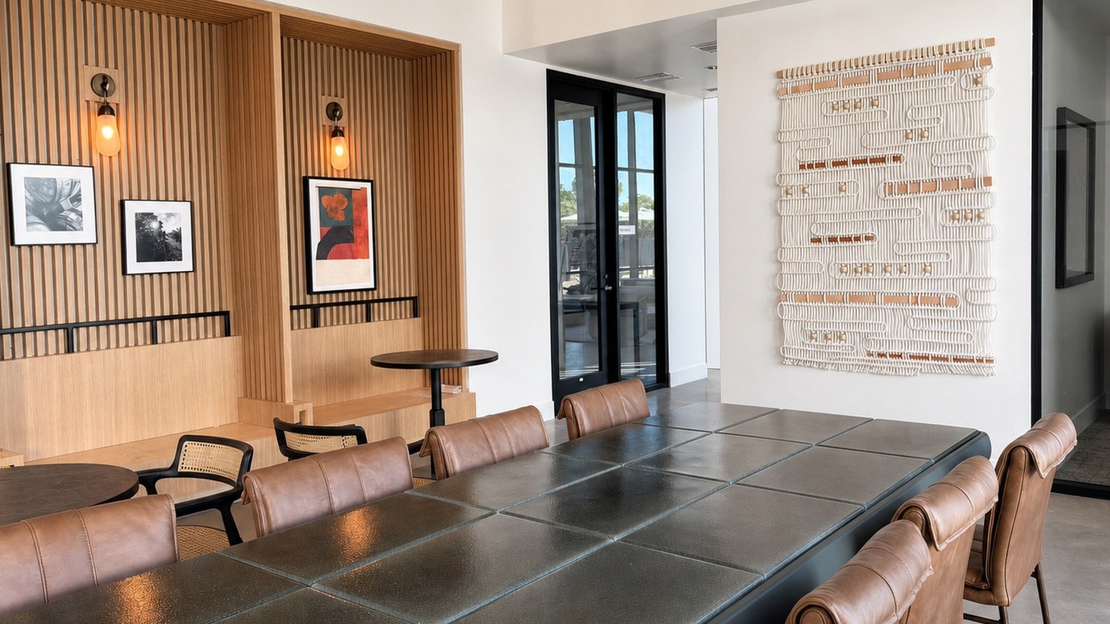 Modern dining area with brown leather chairs around a large black table, wooden wall decor with framed black-and-white and colored artwork, wall-mounted light fixtures, and a textured white art piece on the white wall.