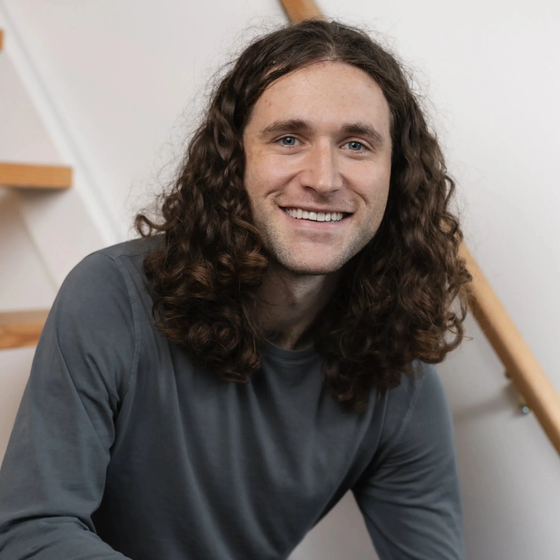 Close-up of a smiling man with long, curly brown hair, wearing a dark gray shirt, sitting indoors against a white wall with wooden stairs in the background.
