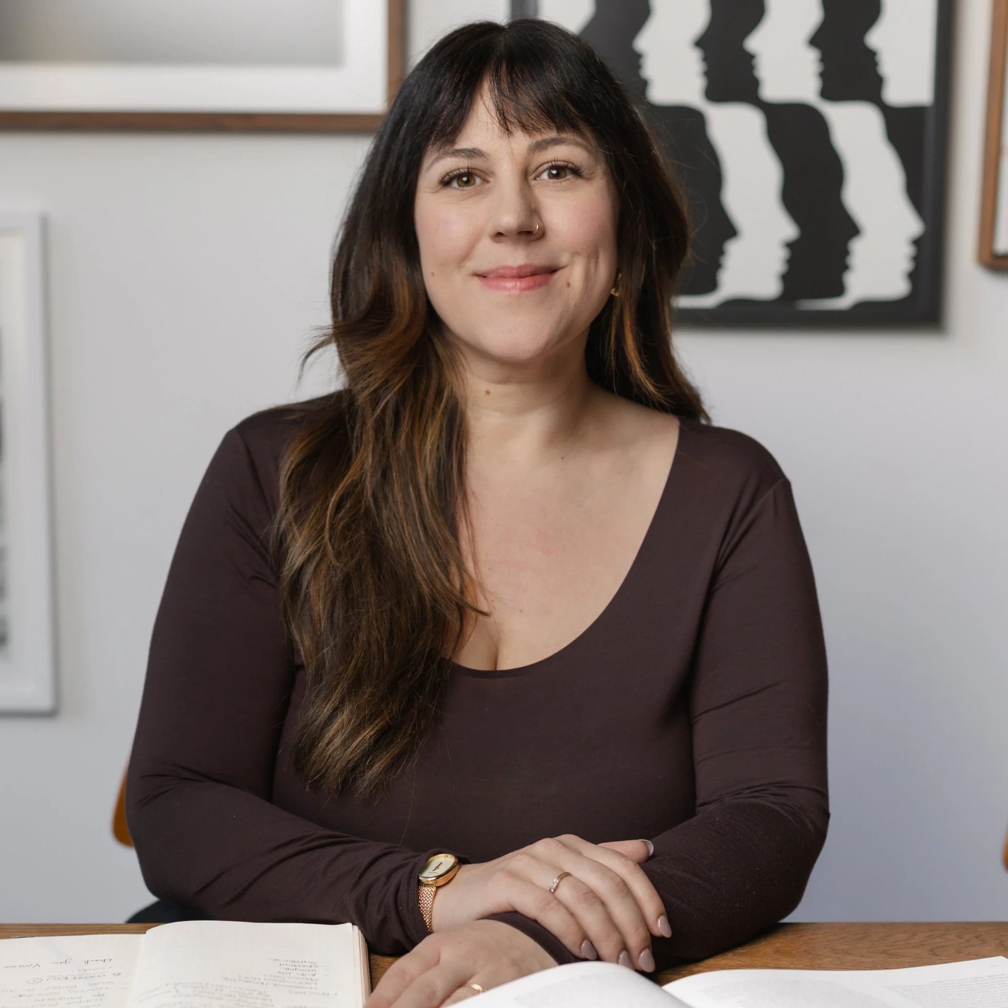 A woman with brown hair and a nose ring sitting at a table with an open notebook, wearing a dark long-sleeve top and a gold watch, smiling slightly, with black artwork featuring silhouettes in the background.