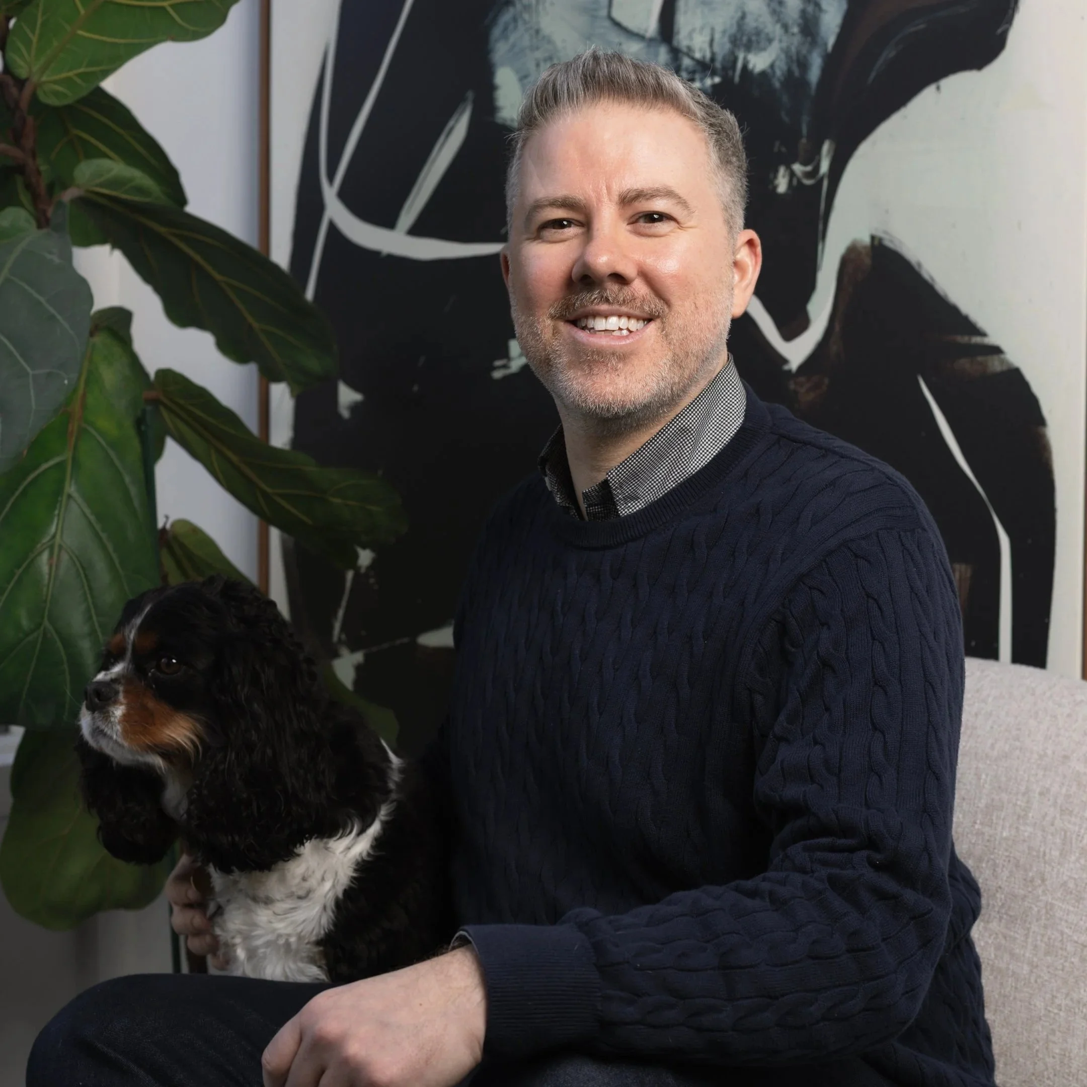 A man with gray hair and a beard smiling, seated indoors near a large leafy plant and abstract black-and-white artwork.