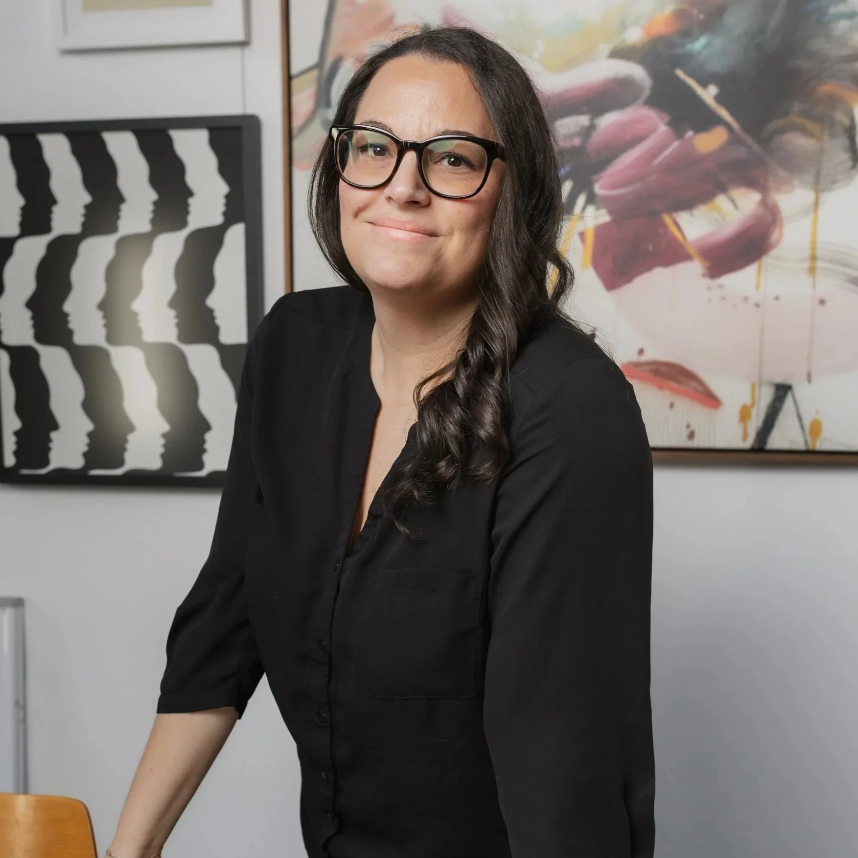 A woman with long dark hair and glasses smiling in an art gallery, with abstract paintings in the background.